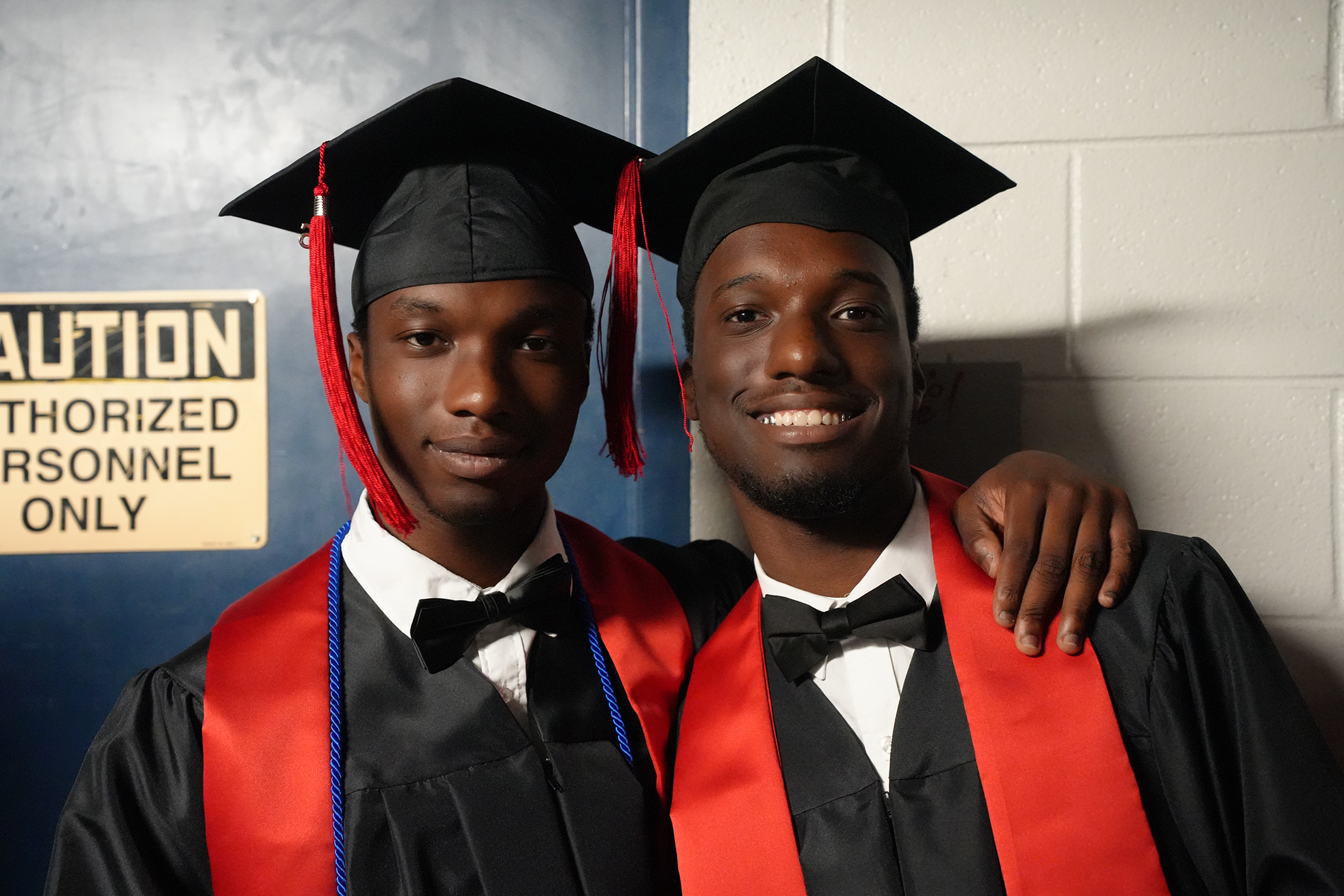 Two men in black and red graduation robes and caps stand together in front of a wall.
