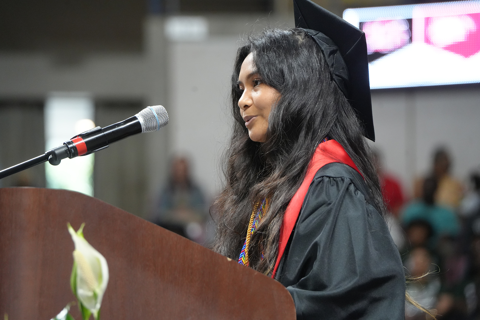 A person in a black graduation gown with a red sash stands at a podium with a microphone.