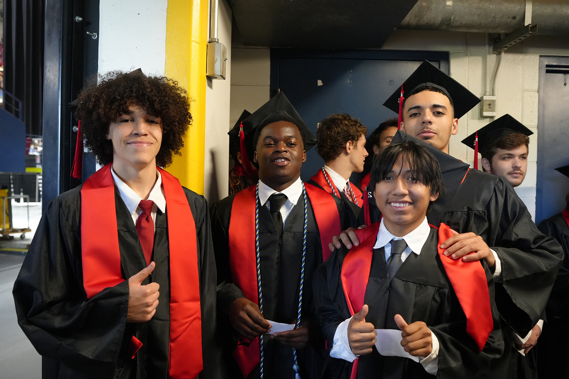 Four young men in black and red graduation gowns, ties, and caps smile and pose for a photo.