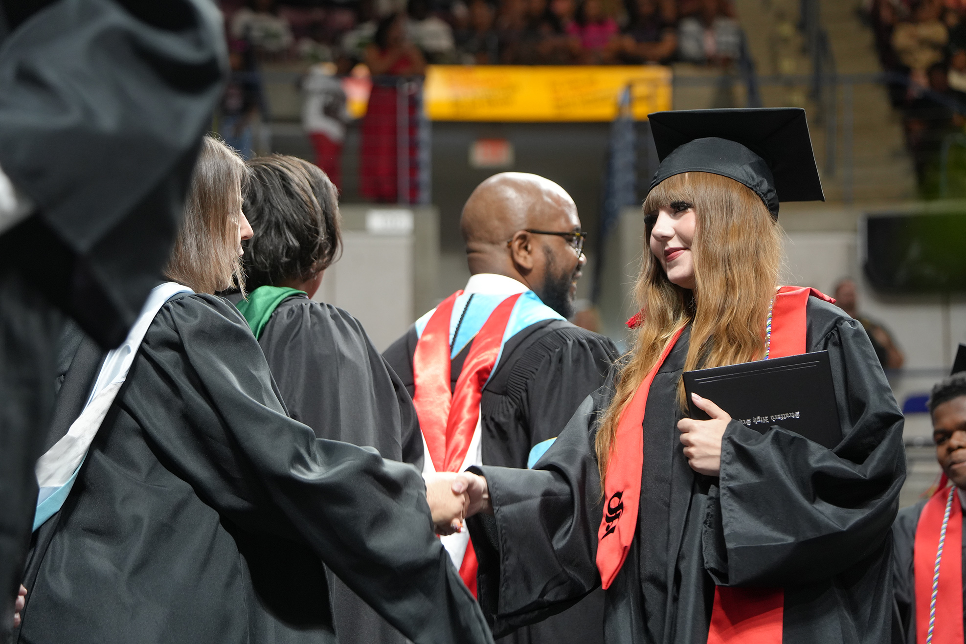 Graduates in academic regalia stand in a line, smiling and shaking hands with one another at a ceremony.
