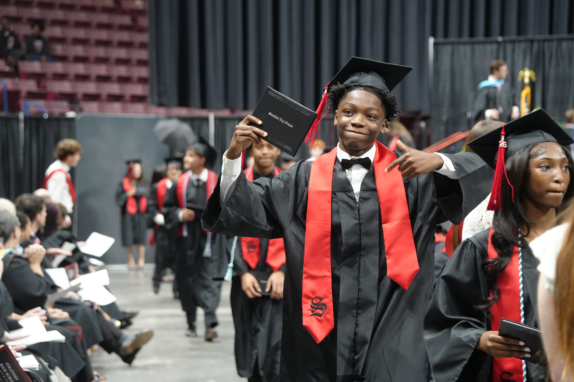 A man in a black cap and gown holds up his diploma. Other graduates stand behind him.