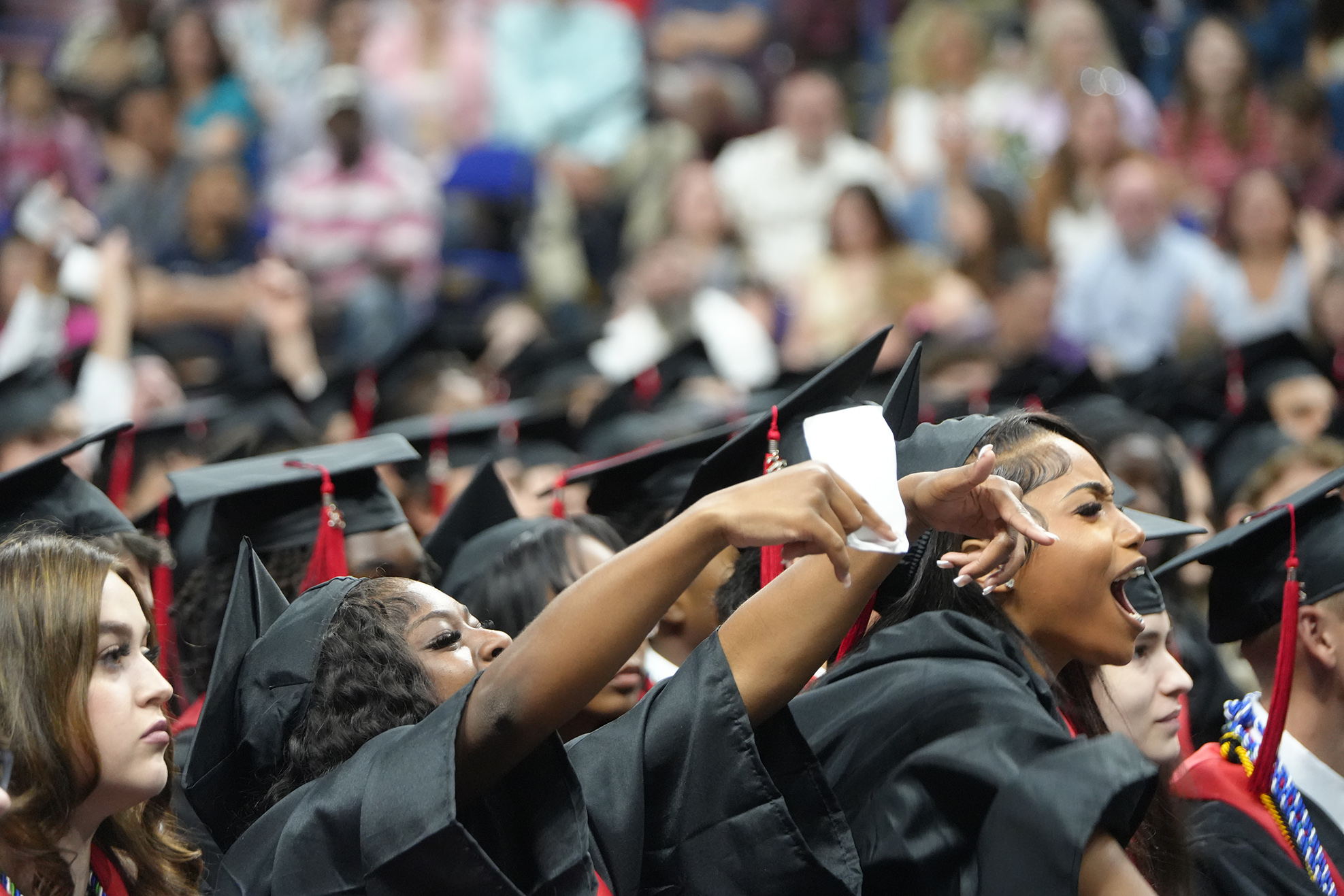 A crowd of graduates in black caps and gowns, some cheering, in an auditorium with many spectators.