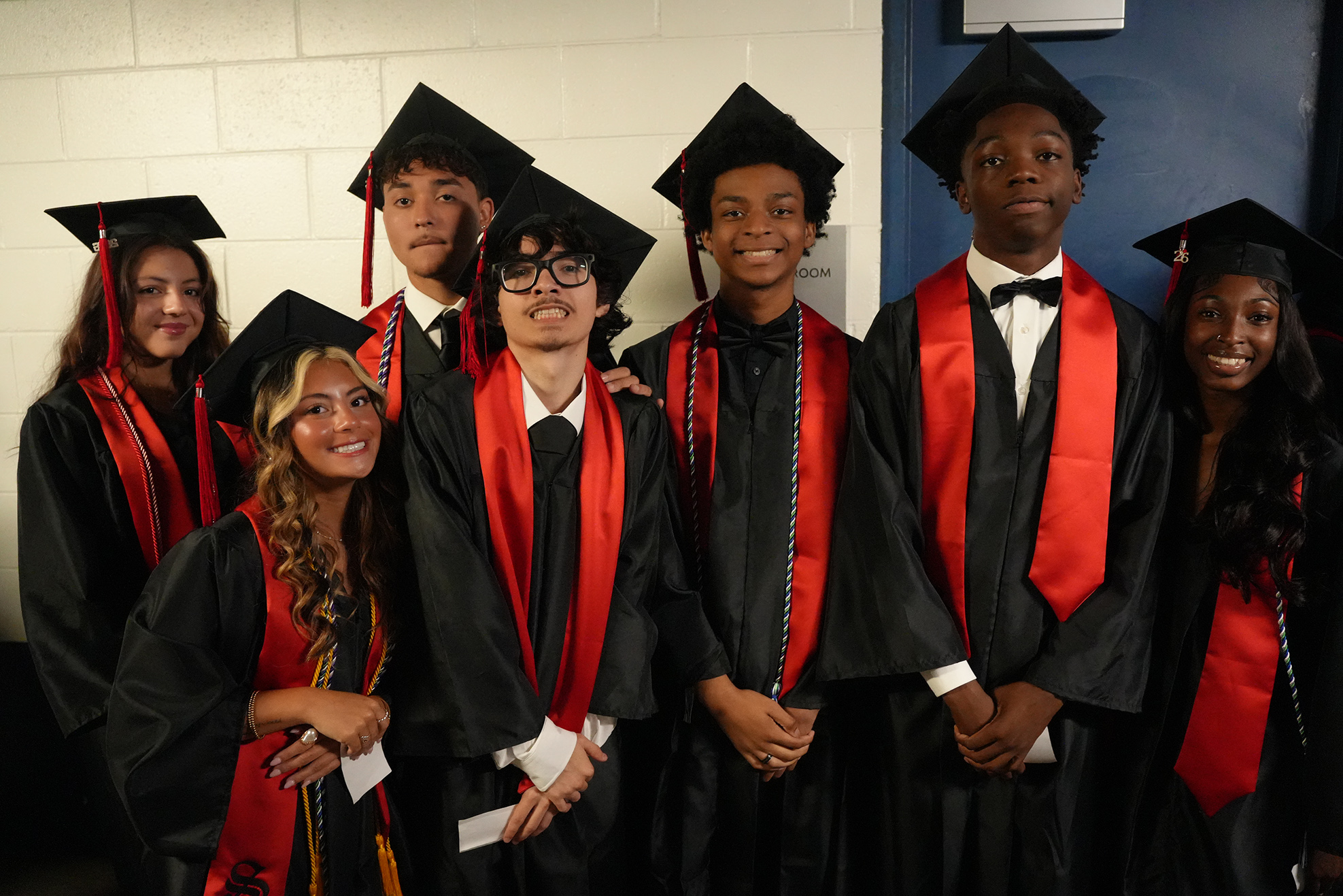 Nine people in graduation attire stand in a row. They are all wearing hats and have sashes over their shoulders.