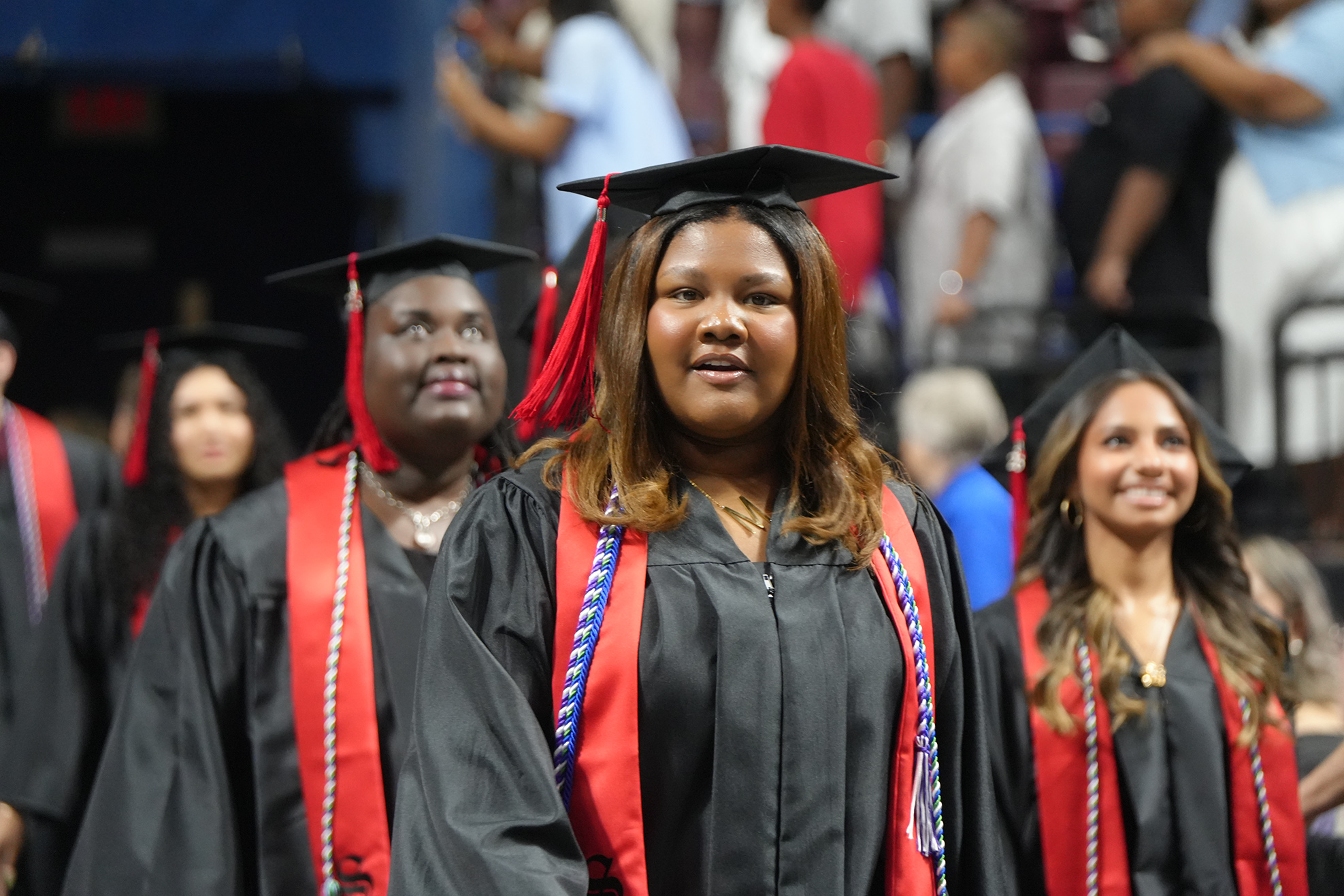 Graduates in black gowns with red sashes and red and black graduation caps walk in a line.