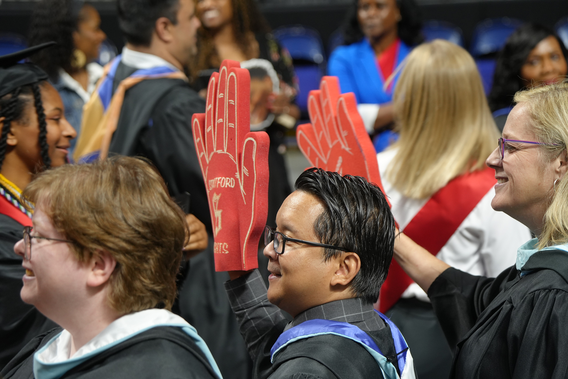 People in academic gowns at a ceremony; a man holds up red gloves while a woman smiles behind him.