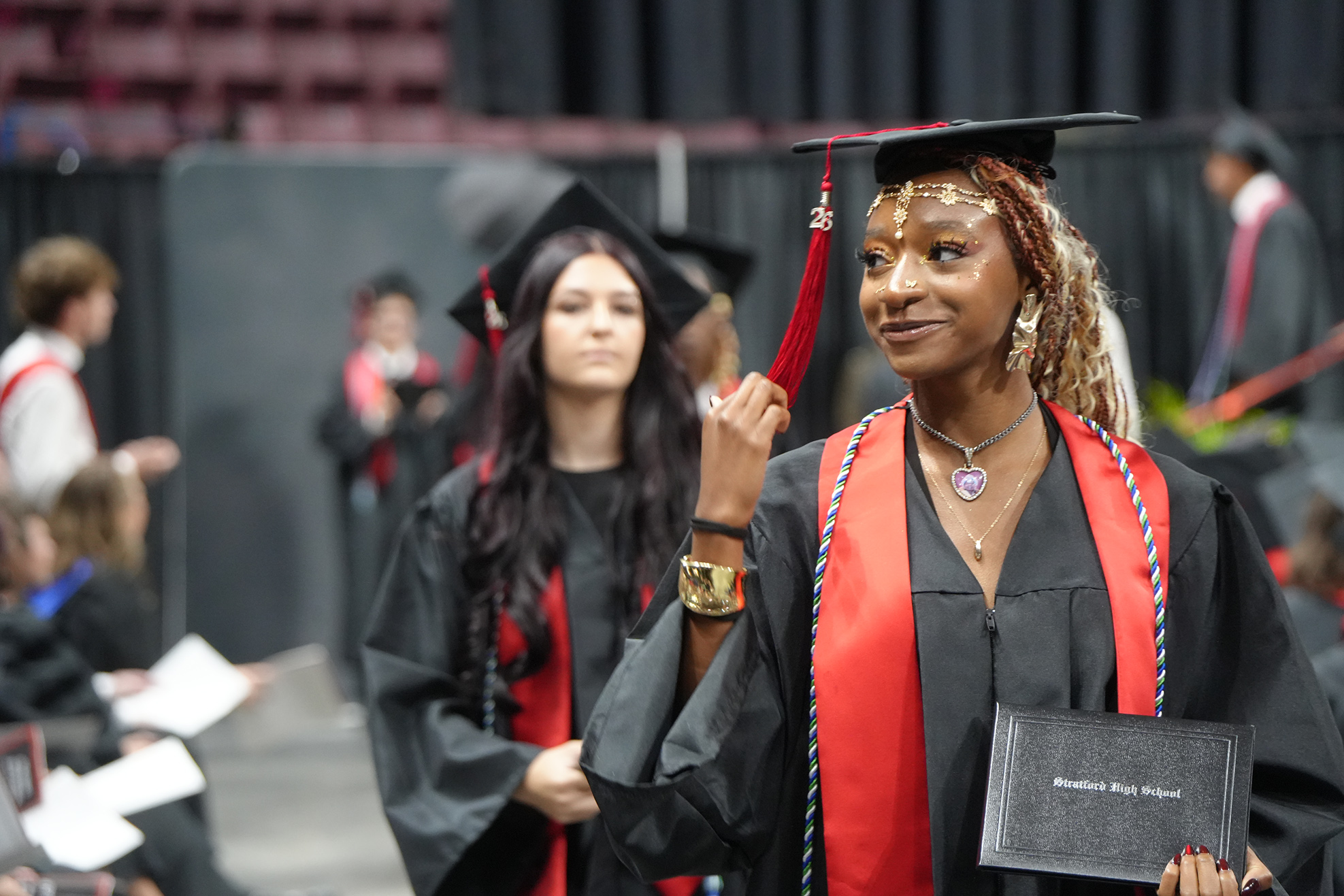 Two women in academic regalia, one holding a diploma, walk in a graduation ceremony. One woman looks ahead.