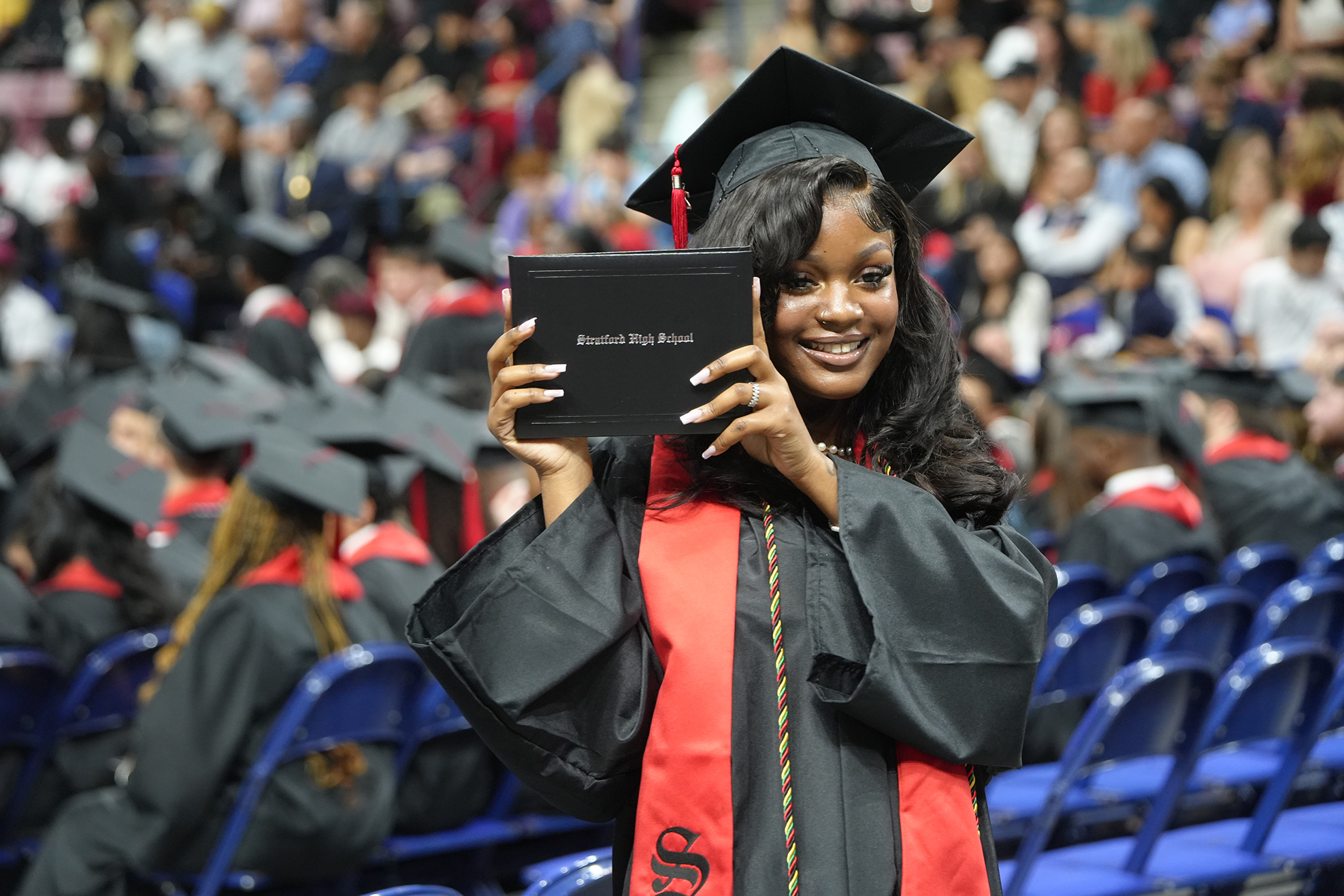 A woman in a cap and gown holding a diploma smiles for the camera at a graduation.