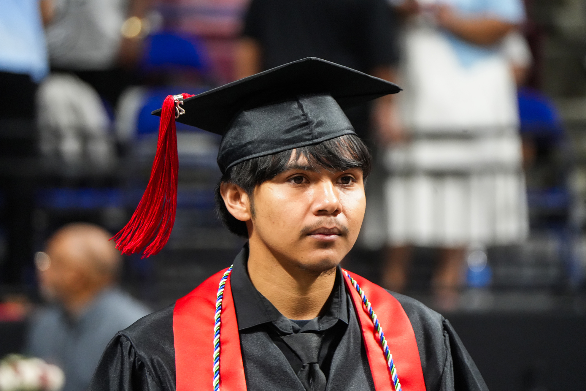 A male graduate stands in a crowd, wearing a black gown and cap with a red tassel.
