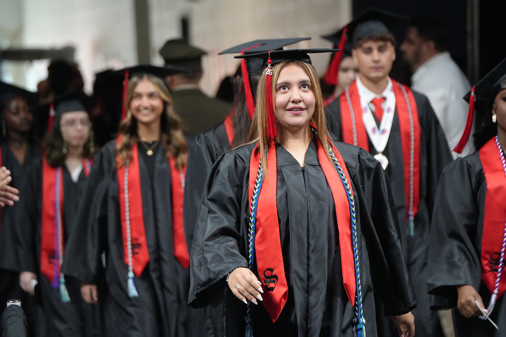 Graduates in black gowns with red sashes and blue tassels walk into an auditorium. Some wear caps.