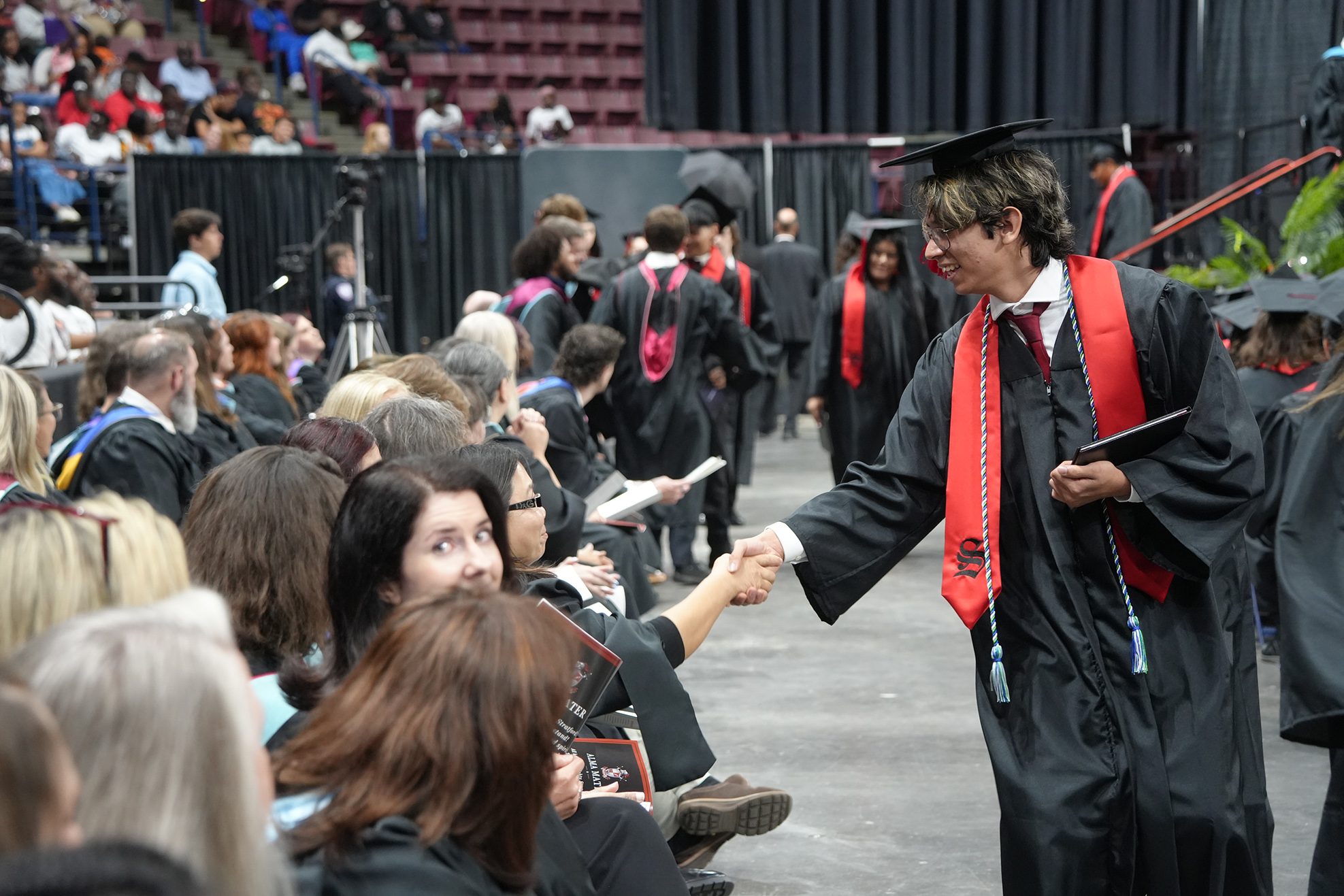 People in caps and gowns, likely at a graduation ceremony, with a graduate shaking hands with a person in front of him.