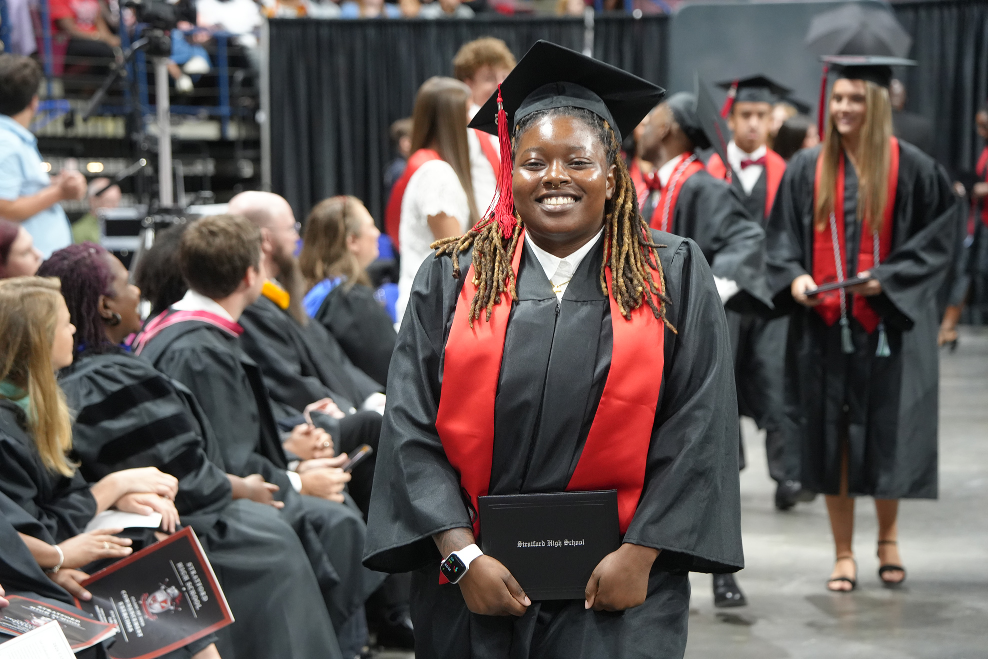 Graduates in black gowns and red stoles sit and stand in a room. A smiling graduate in the foreground holds a diploma.