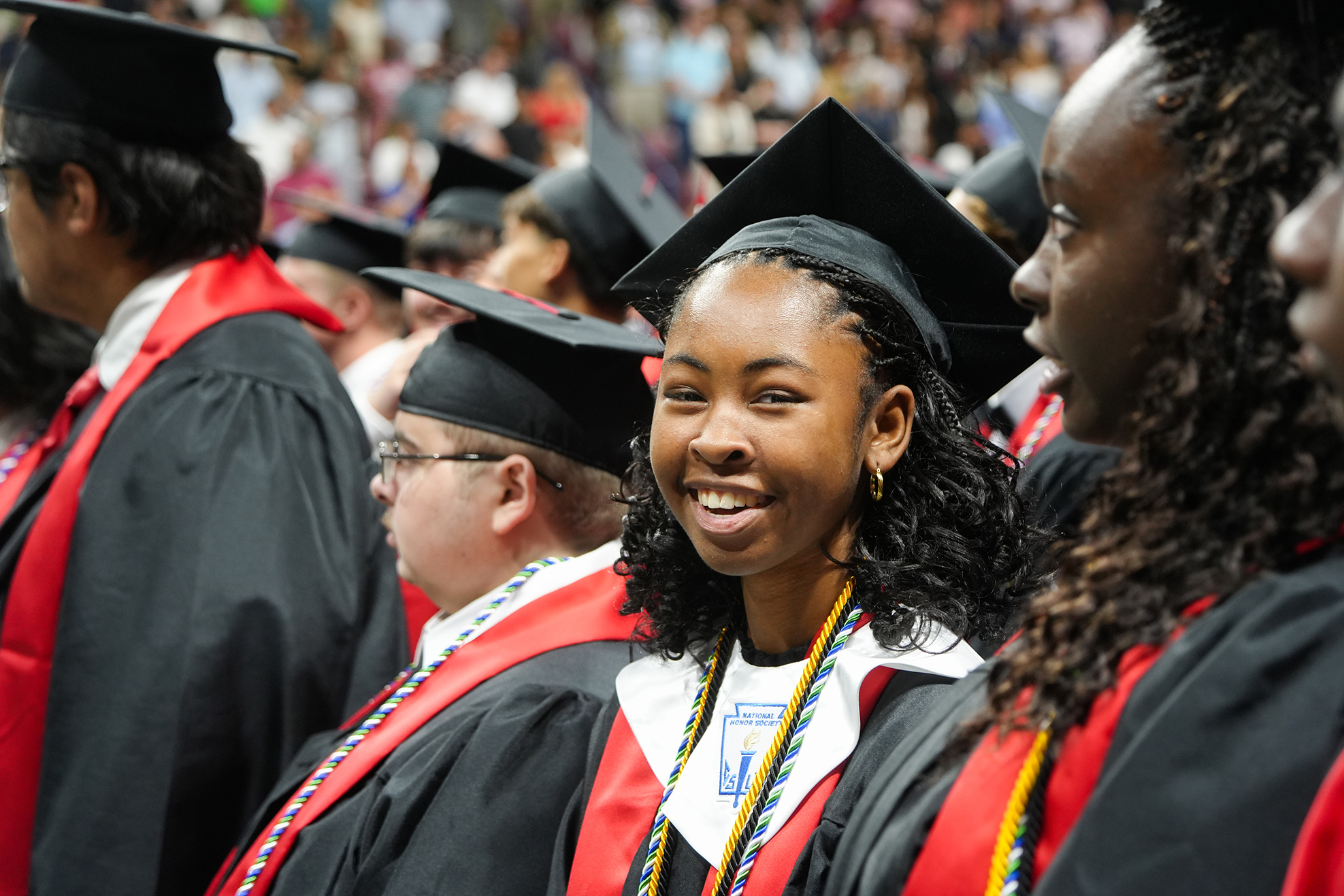 Graduates in black caps and gowns smile, stand, and wait. Some wear glasses and colored cords.