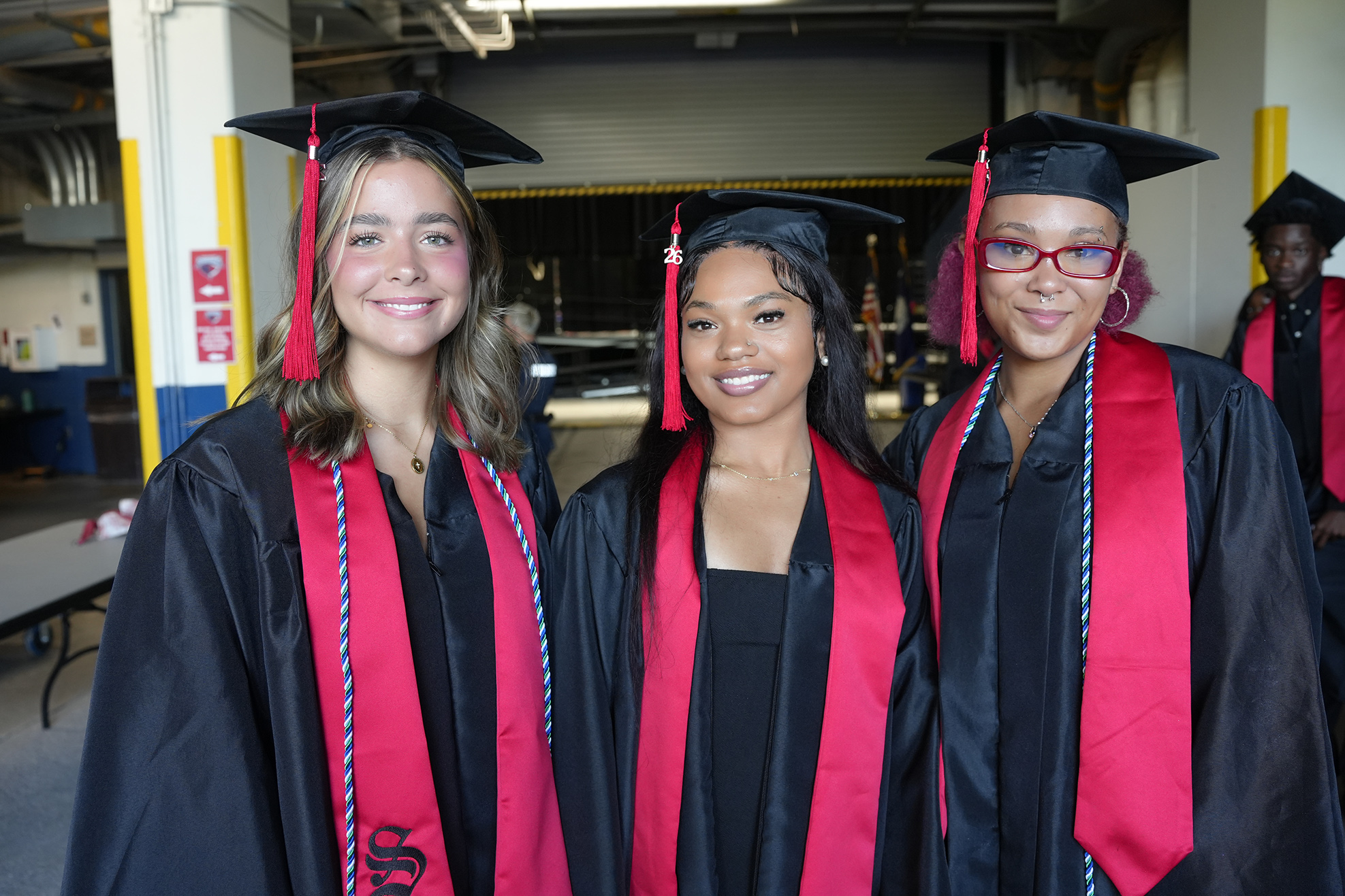 Three women in black and red gowns with tasseled caps pose for a photo in a room.