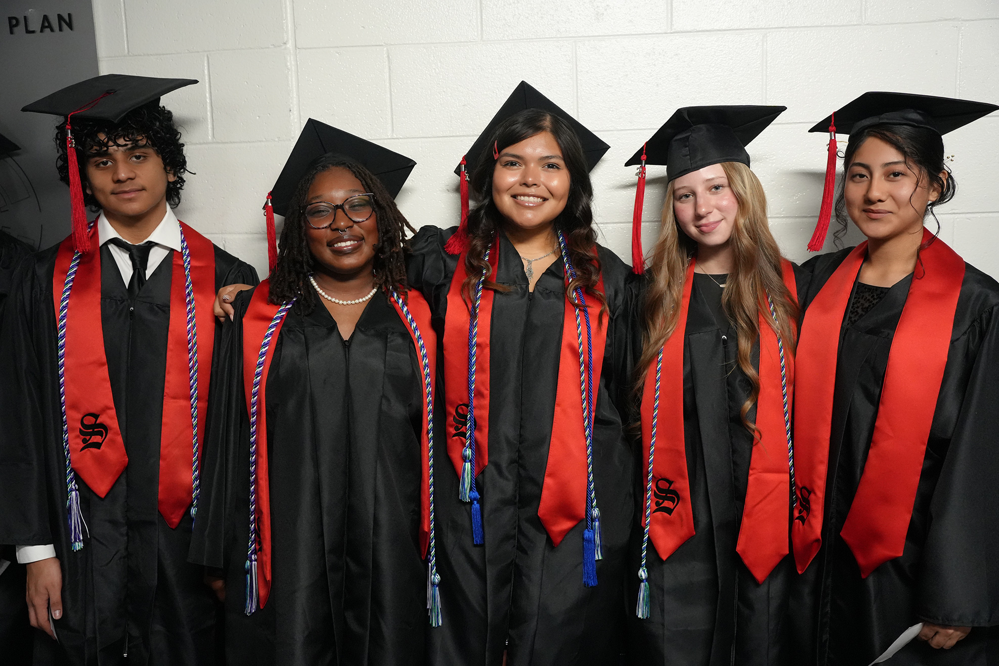 A group of six young adults wearing graduation gowns, caps, and stoles stand together.