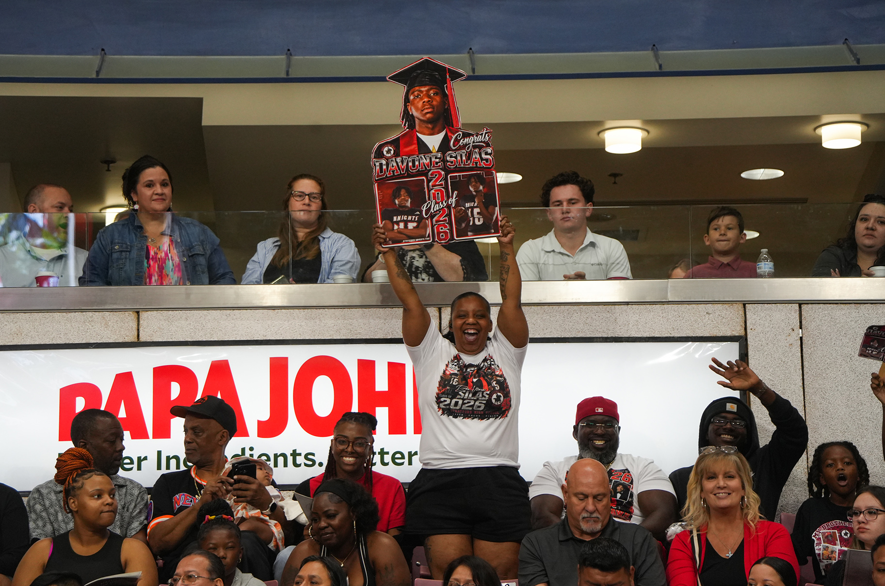 Audience members at a sporting event, some holding up signs, and a banner for Papa John's.