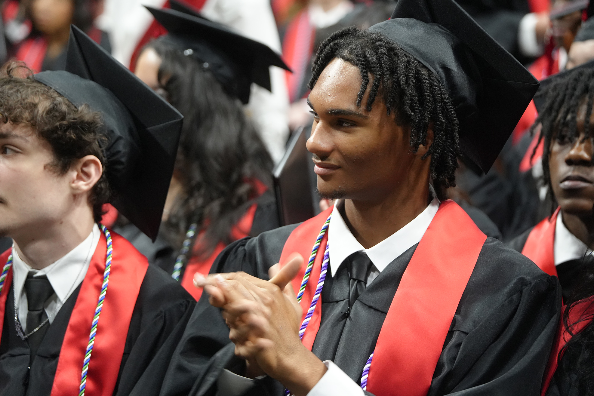 Several people in black graduation gowns and caps, seated, with a focus on a man clapping.