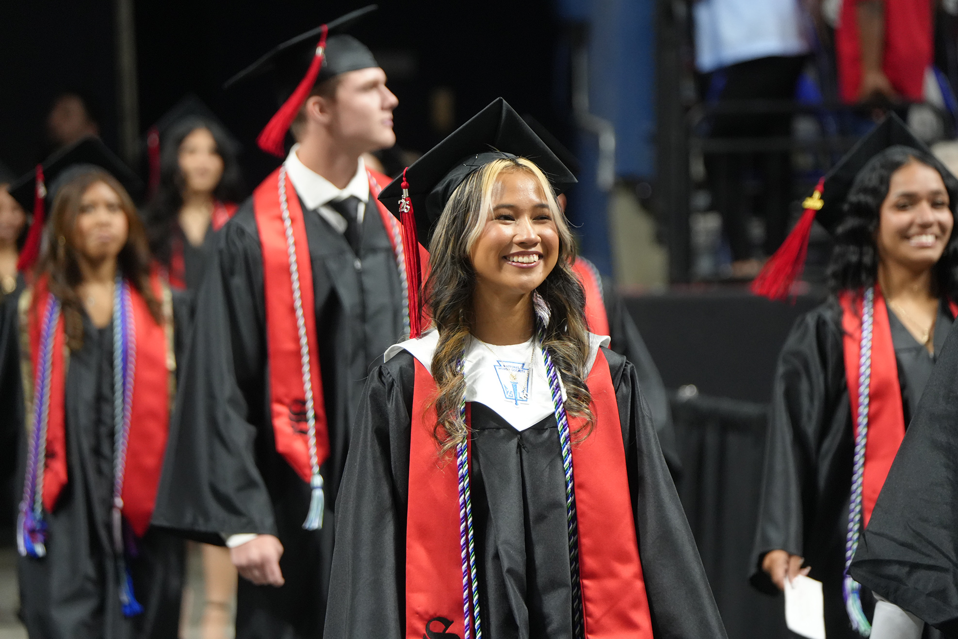 Graduates in black and red gowns and caps walk together in a line at an outdoor graduation ceremony.