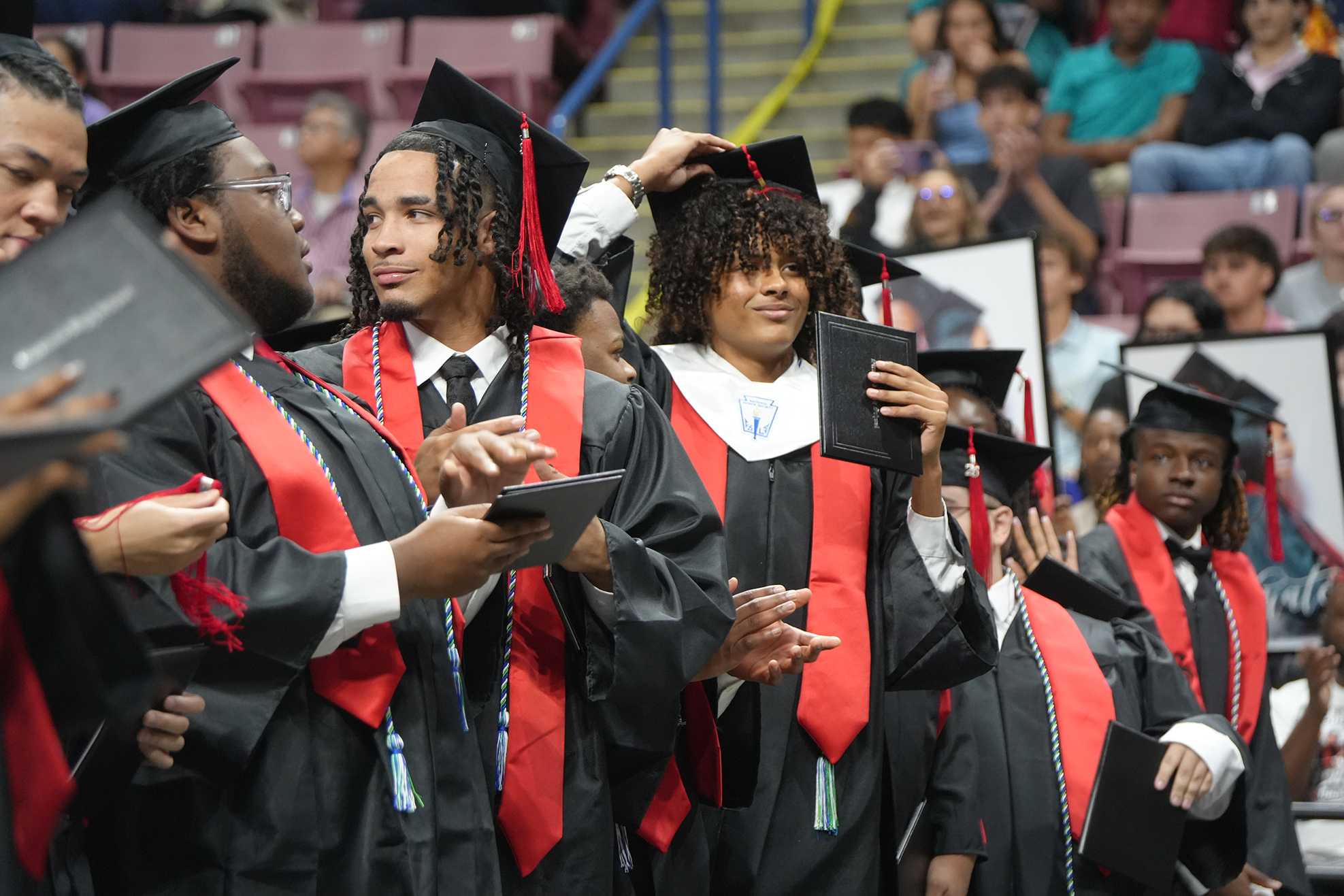 Graduates in caps and gowns, standing in a row at a ceremony, some holding diplomas.