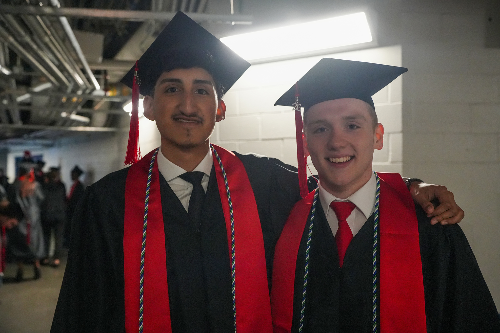 Two men in graduation robes with red sashes and mortarboard hats standing close to each other.