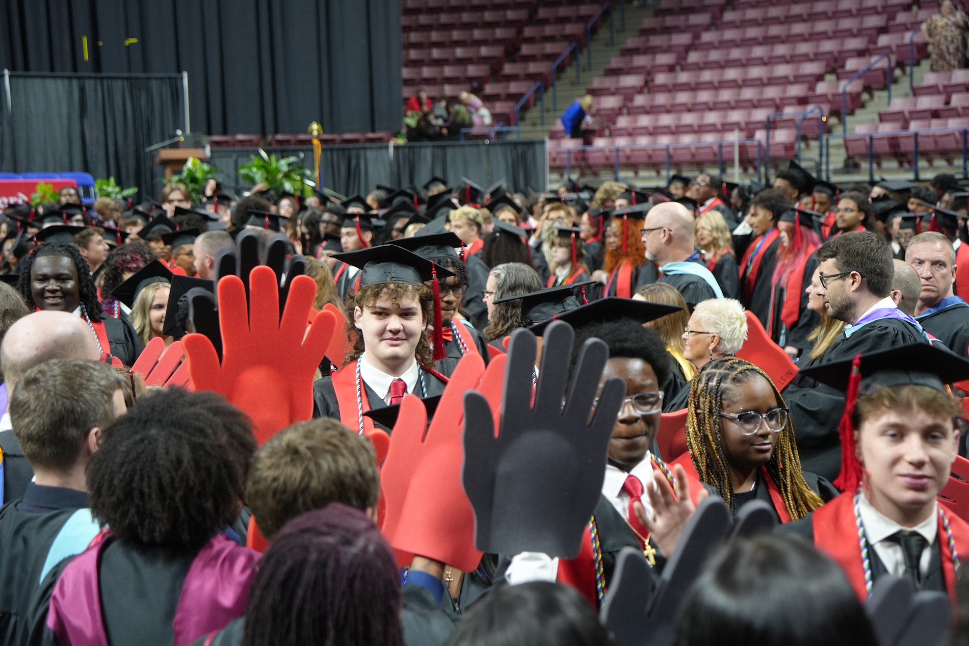 A crowd of people in graduation attire, some holding black and red hand cutouts, stand in an arena.