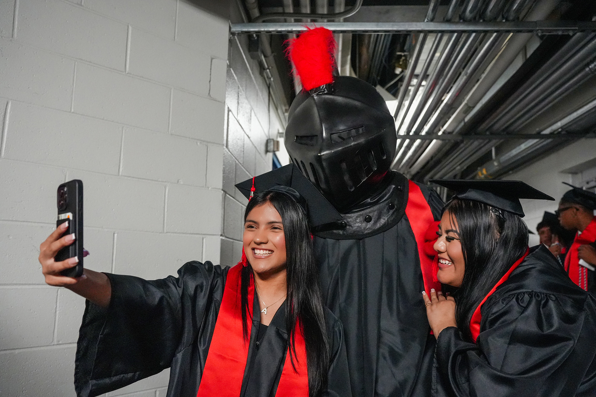 Two women in black and red gowns and a person in a black costume stand against a white wall, one taking a selfie.