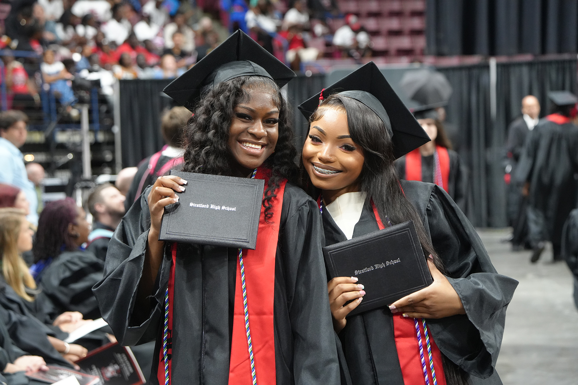 Two women in caps and gowns, holding diplomas, stand side by side at a graduation ceremony.