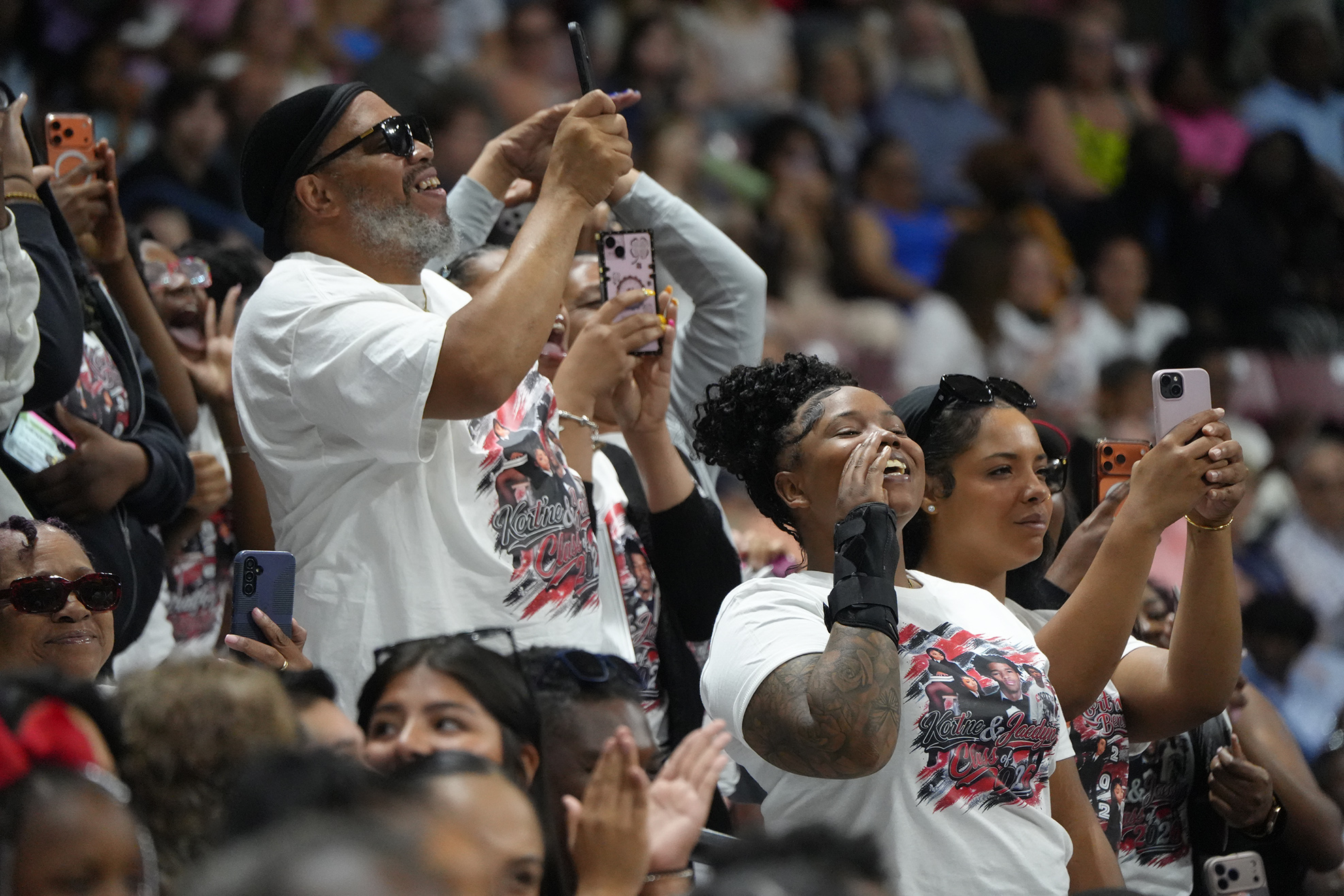 An audience takes photos and videos with their phones, some clap, during an event in a stadium.
