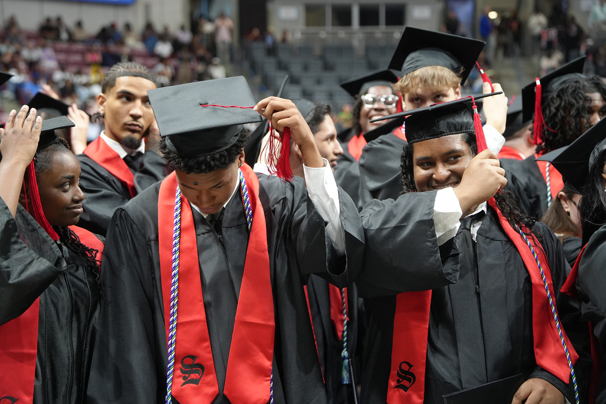 Graduates in black gowns and red stoles stand in a stadium, adjusting hats. Some hold diplomas.