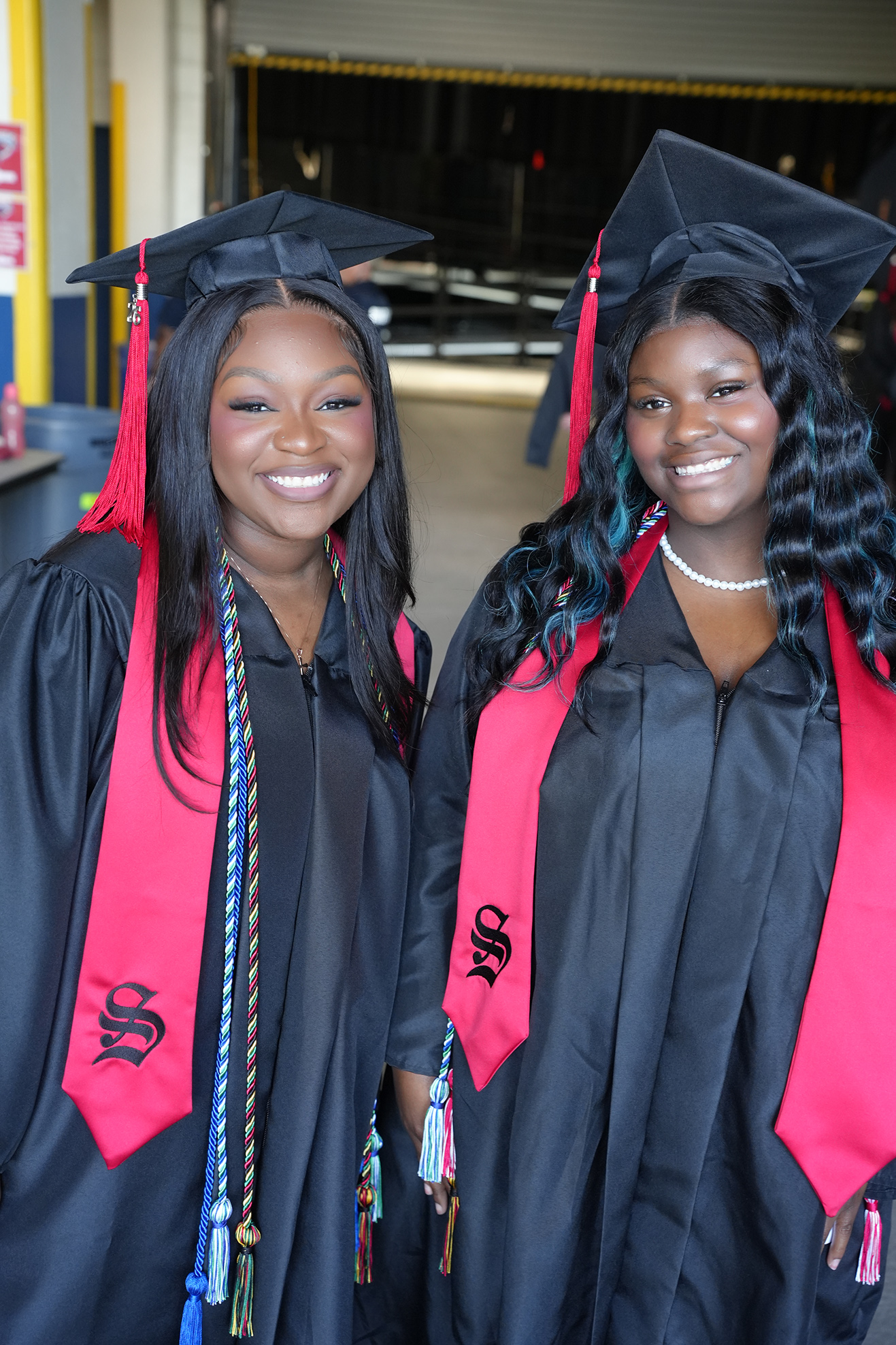 Two women in black and red graduation robes and caps, one with a tassel and the other with a blue-dyed braid, stand together and smile.