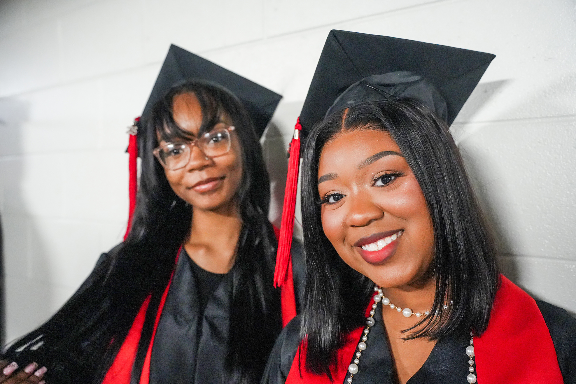 Two women in black and red graduation gowns and caps, one smiling, the other looking forward.