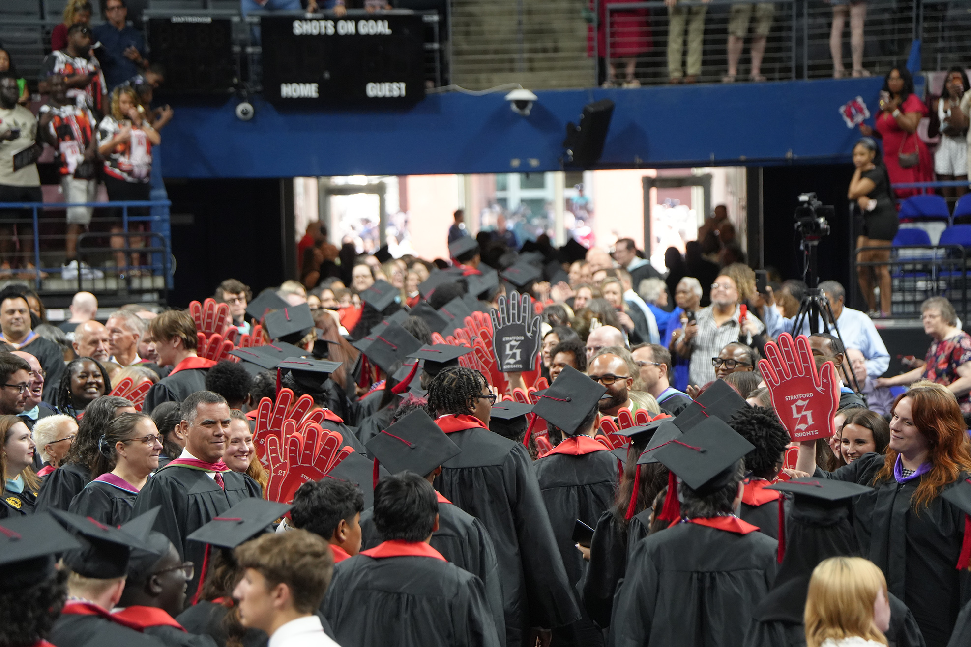 A large crowd gathers indoors for a graduation ceremony. Many people wear black caps and gowns.