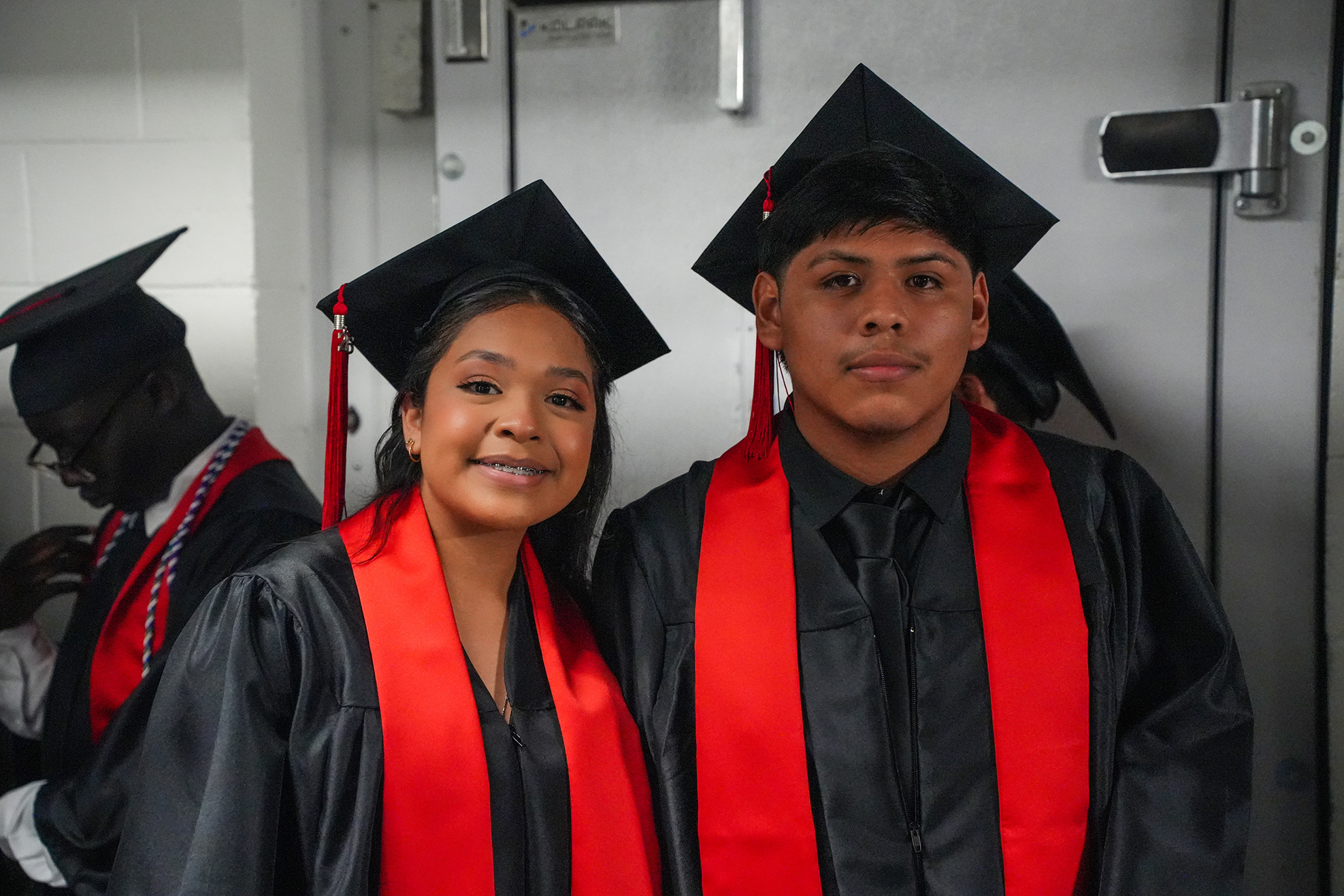 Two individuals in graduation gowns with red sashes, one with a smile and the other with a neutral expression.