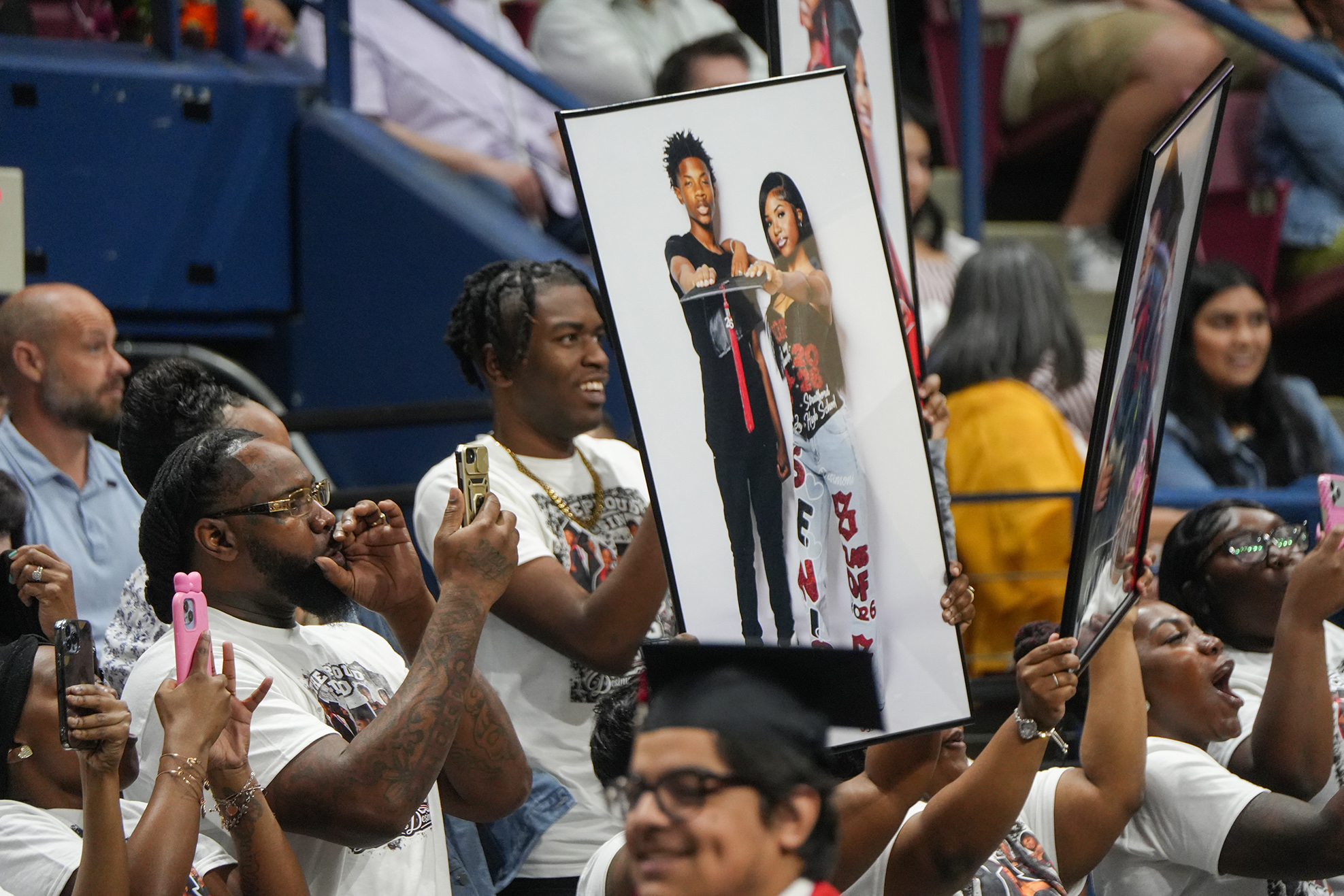 Audience members at a graduation ceremony hold framed photos of graduates, some wearing caps and glasses.