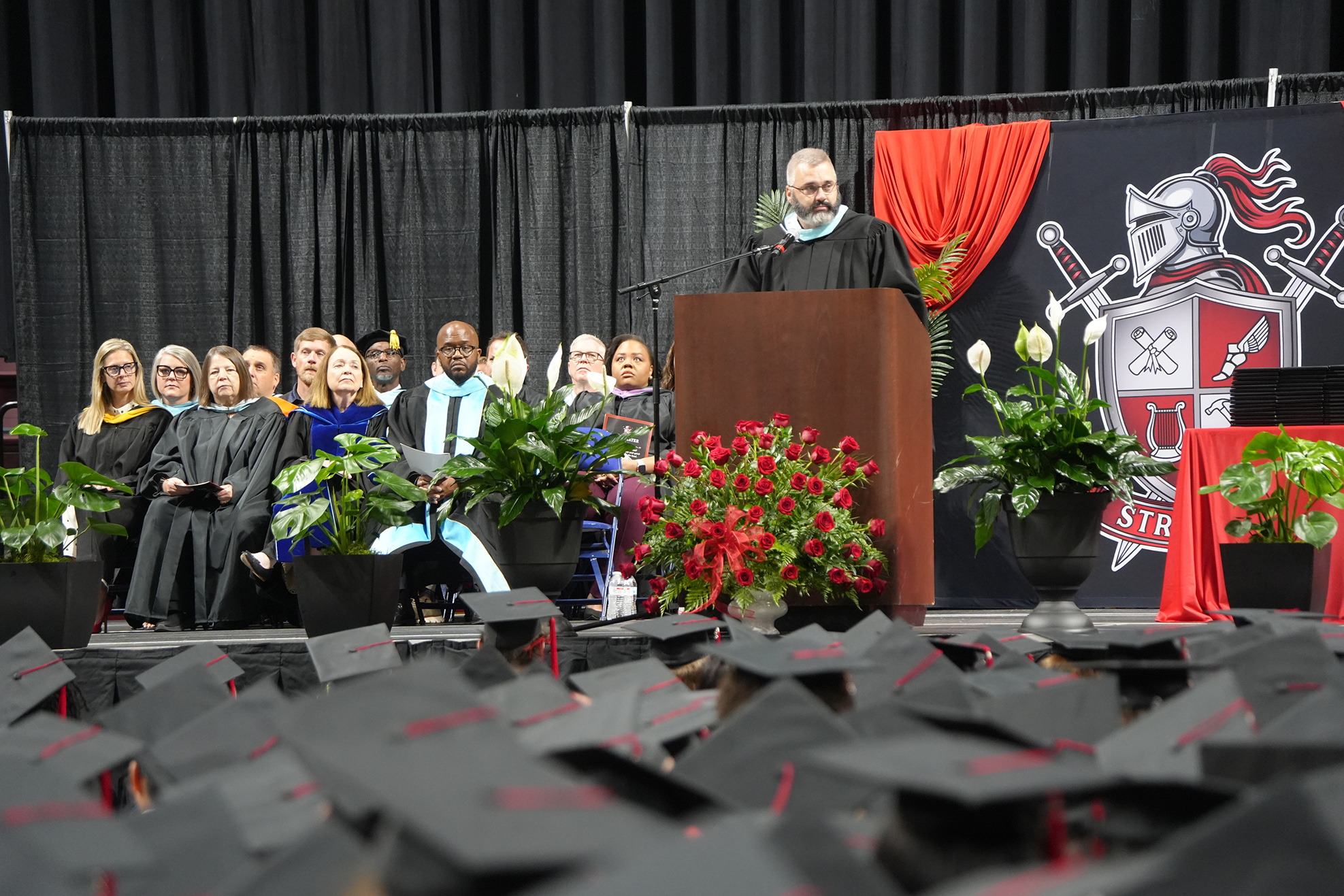 A speaker at a podium during a graduation ceremony, with a stage backdrop and seated audience in caps and gowns.