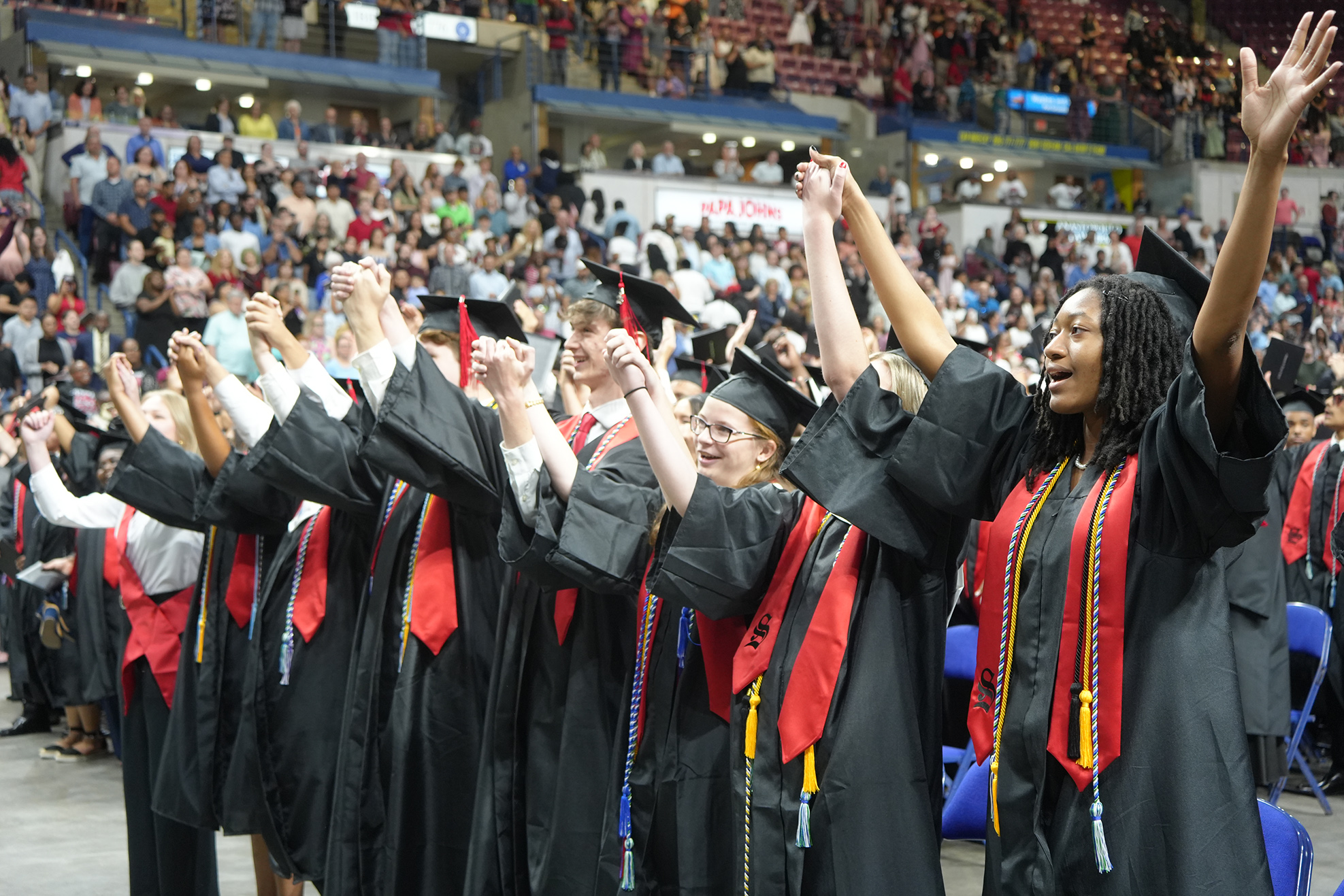Graduates in black gowns and red stoles raising their hands in an arena with a large audience.