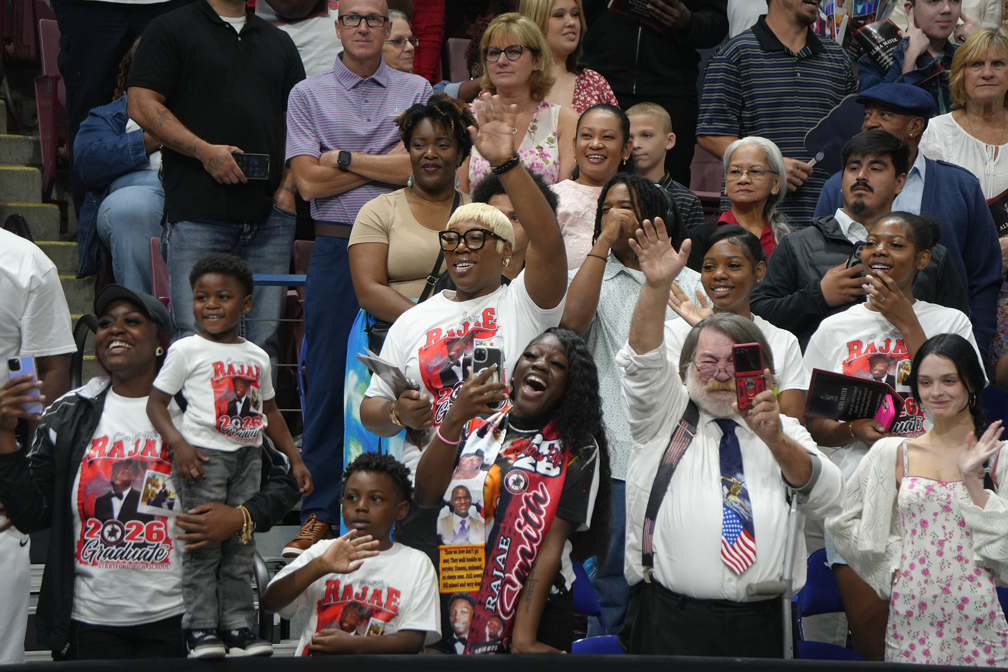 A crowd of people, some wearing white shirts with printed designs, standing and sitting on bleachers, raising hands.