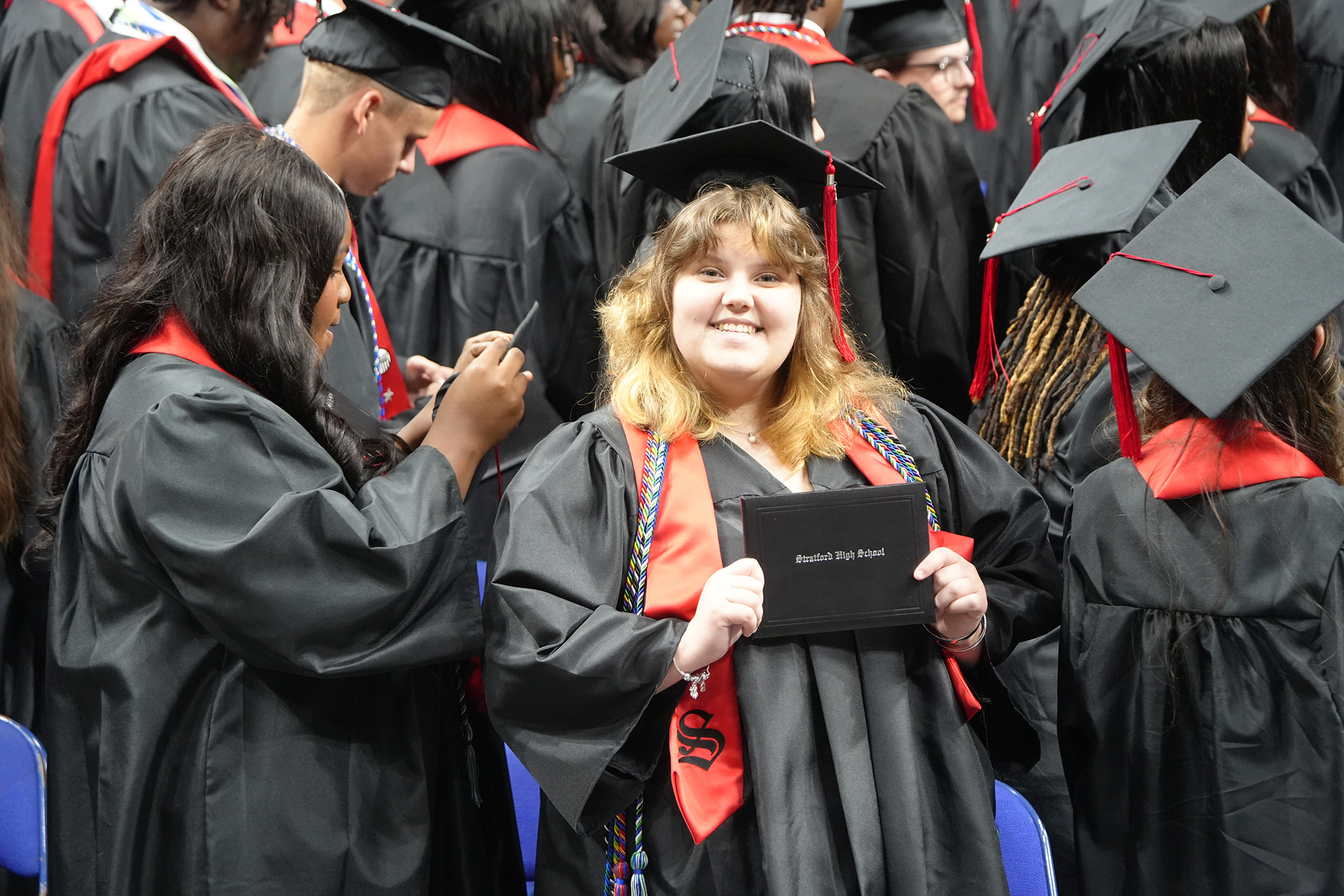 People in black graduation robes and hats stand in a crowd. A woman holds a diploma.