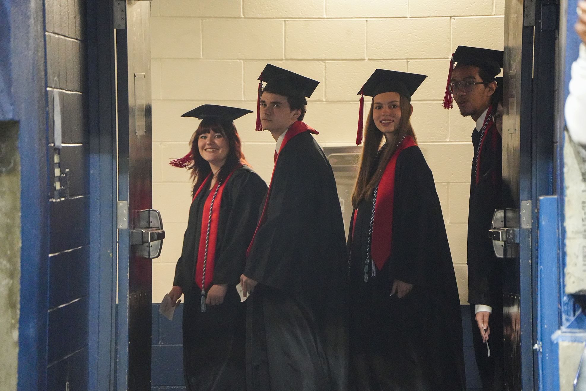 Four individuals in black graduation gowns with red sashes, wearing mortarboard hats, stand in an open doorway.