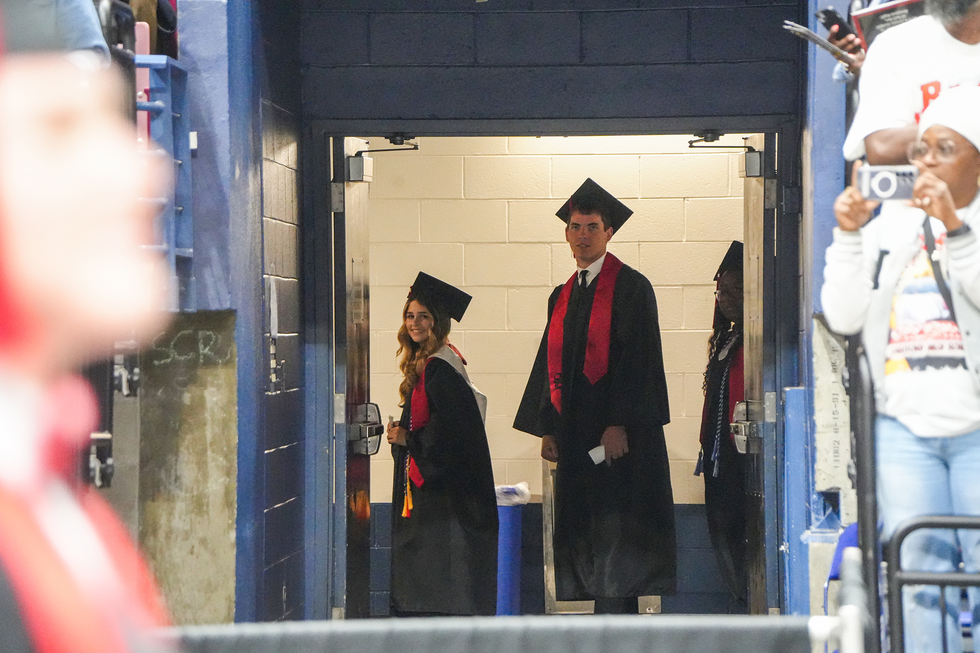 People wearing academic gowns and caps walk through a stadium tunnel; one holds a microphone.