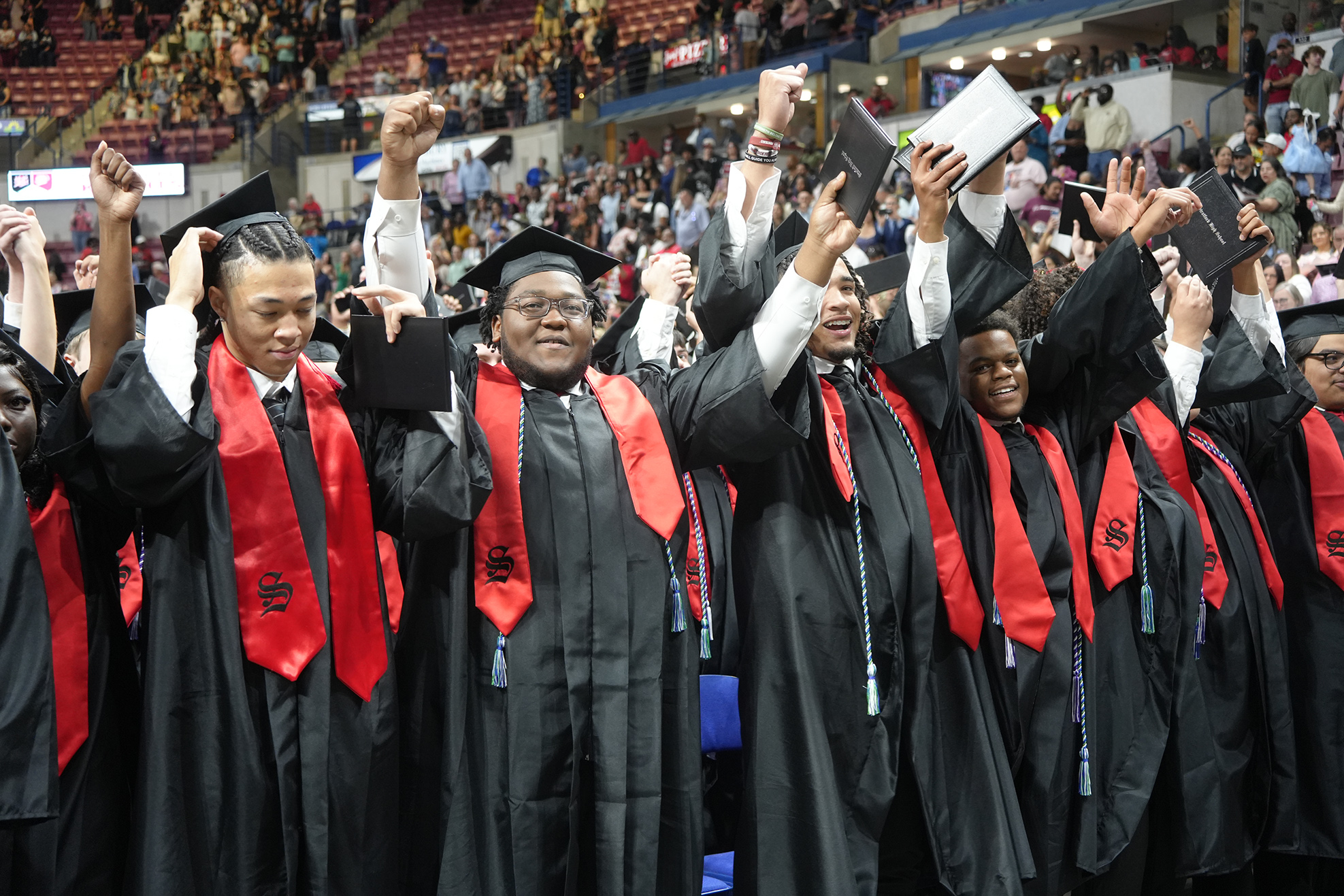 A group of graduates in black gowns with red sashes, raising their hands and caps in a stadium.