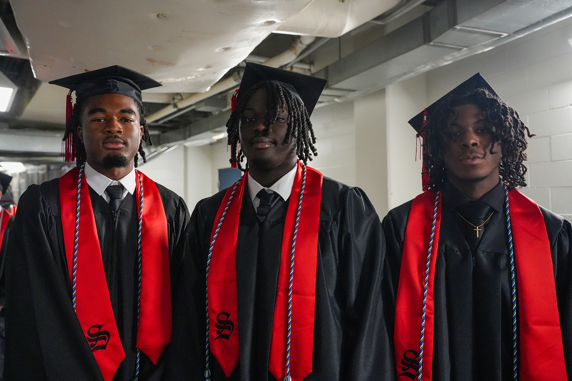 Three men in black graduation gowns and red stoles stand in a hallway, likely at a graduation event.