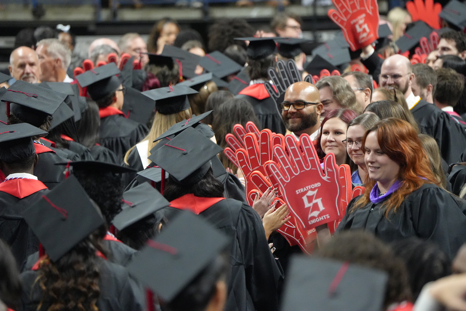 A crowd of people at a graduation, all wearing black caps and gowns, some holding red gloves.