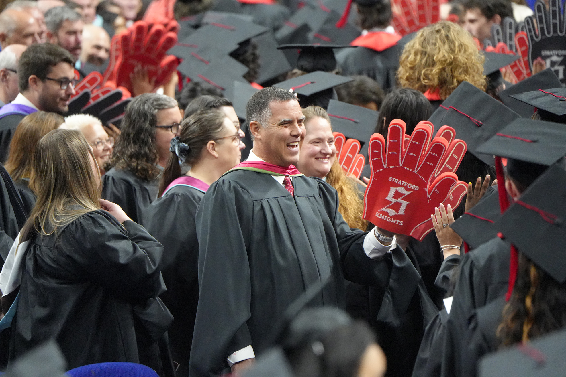 A large crowd of graduates in black gowns and caps at a ceremony; one holds a red sign.