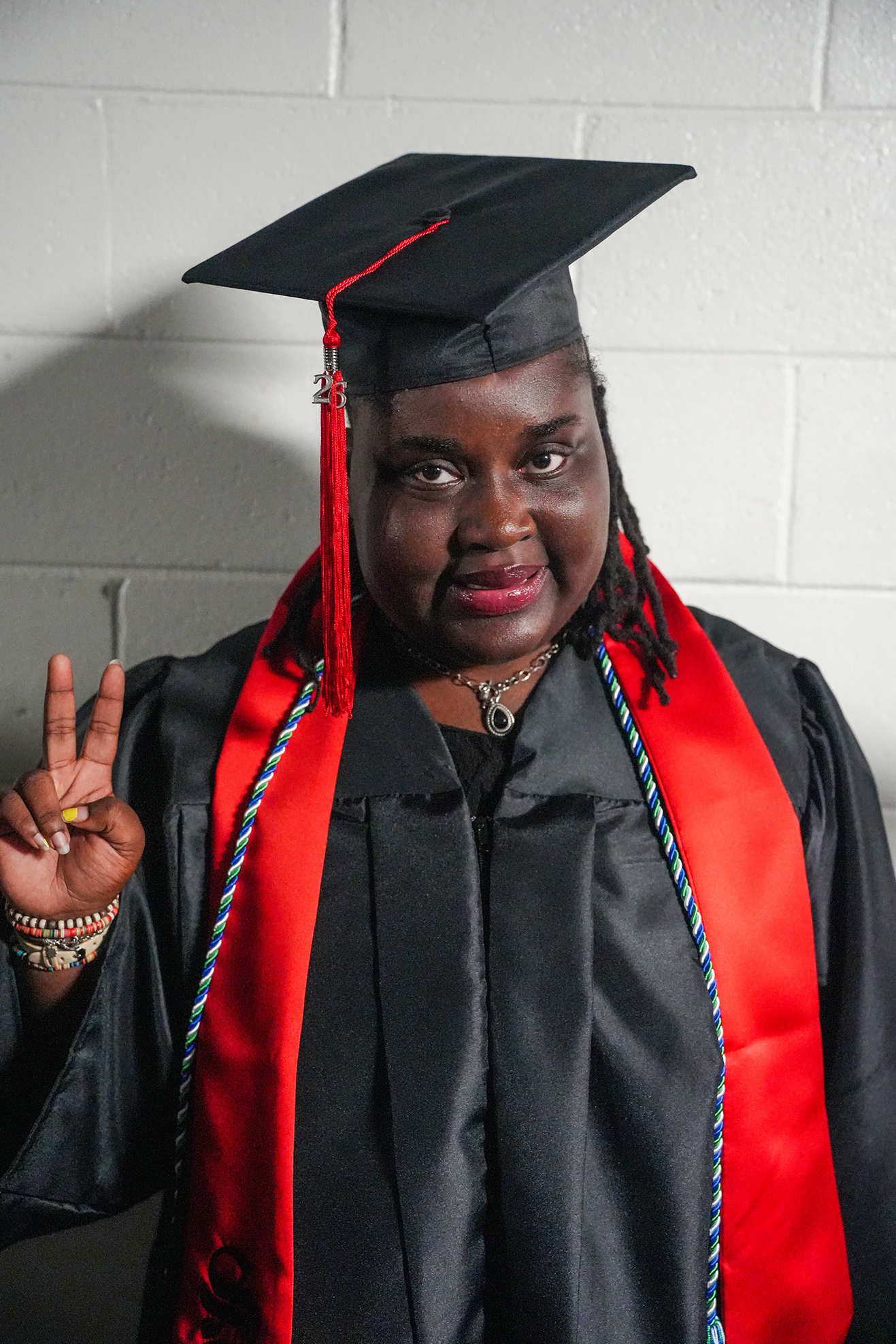 Person in a black graduation gown with red and blue cords poses with a peace sign in front of a brick wall.
