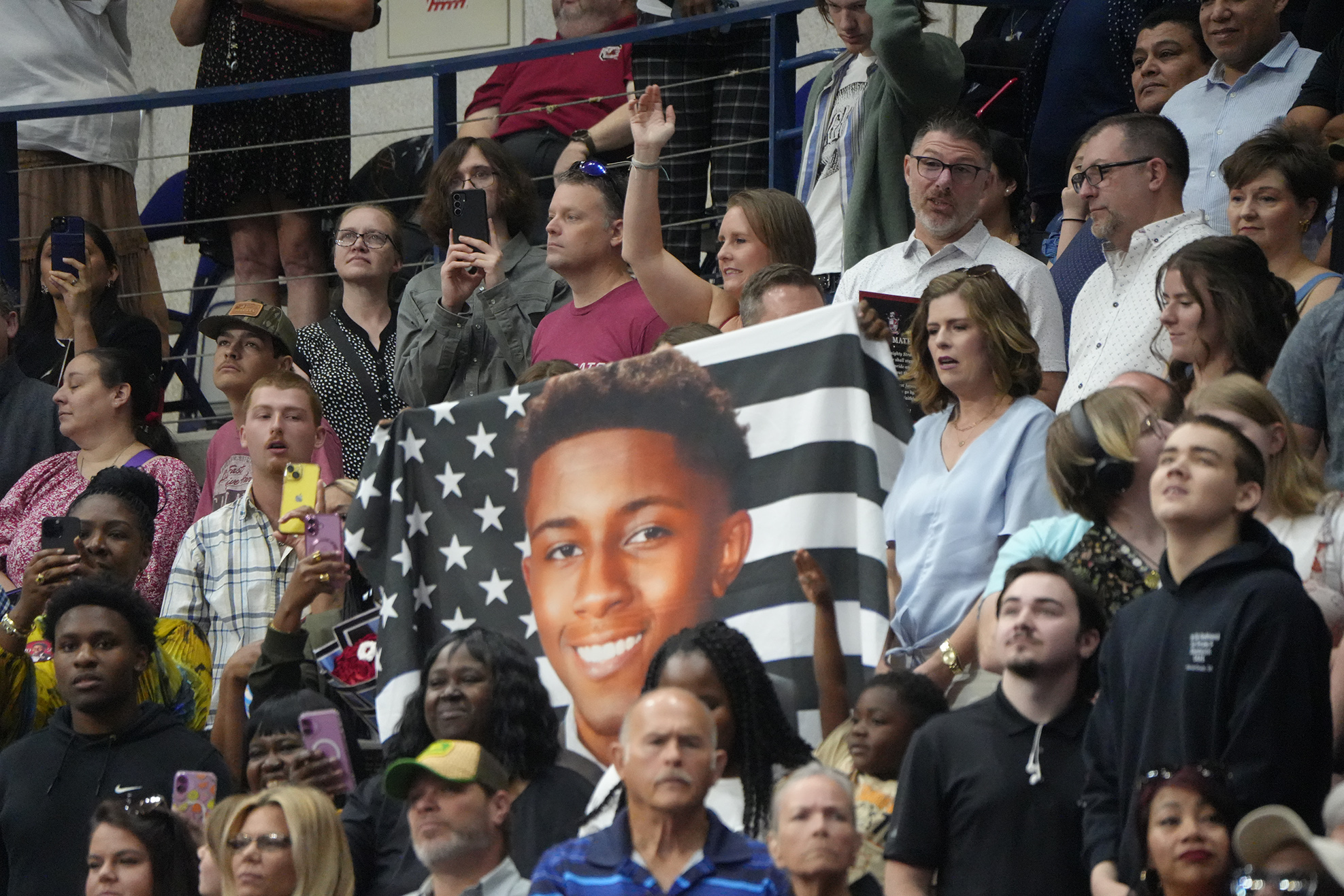 Many people in a stadium hold an American flag with a photo of a smiling man in front.