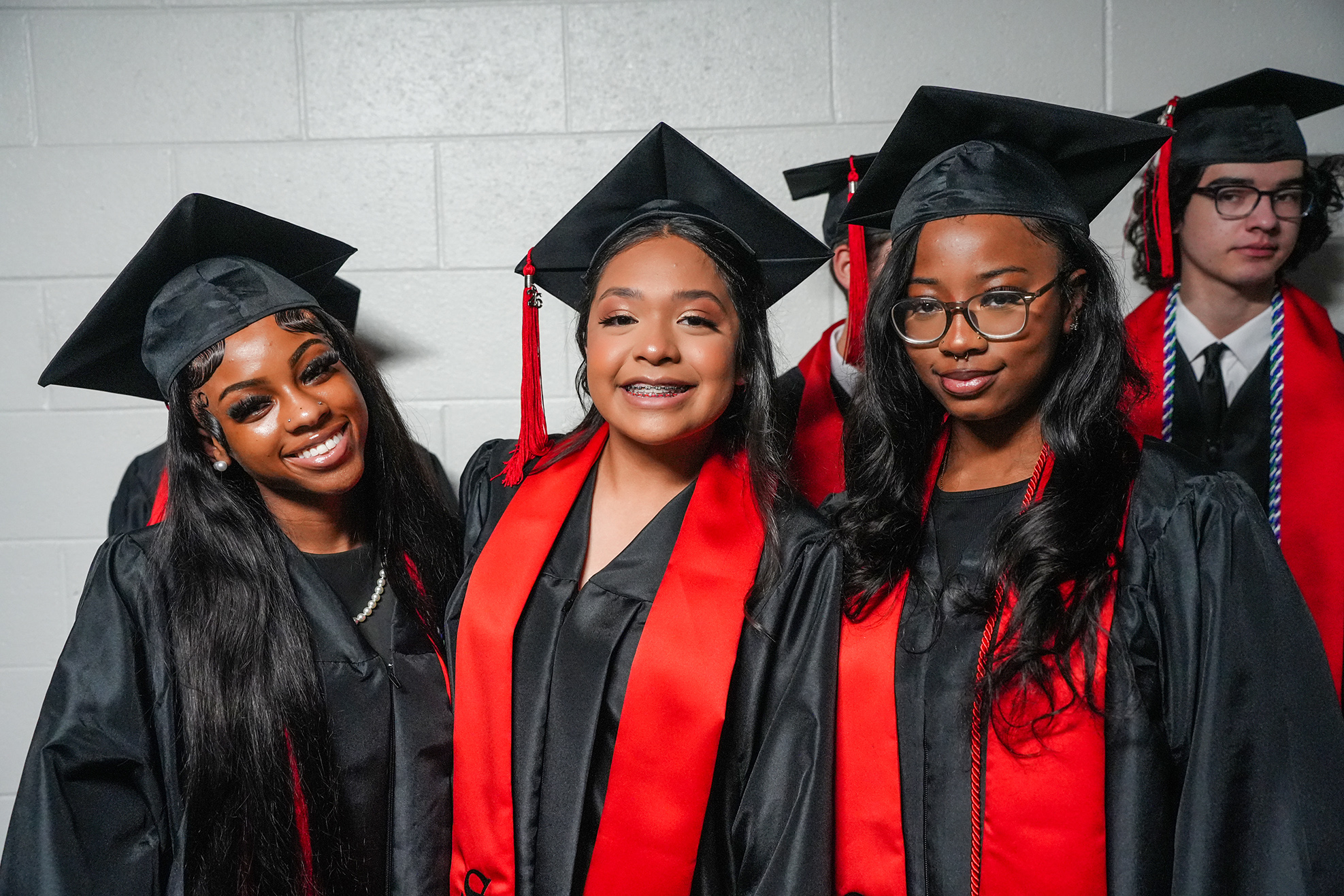 Four individuals in black graduation gowns and caps, two with red stoles, stand in a row against a white brick wall.