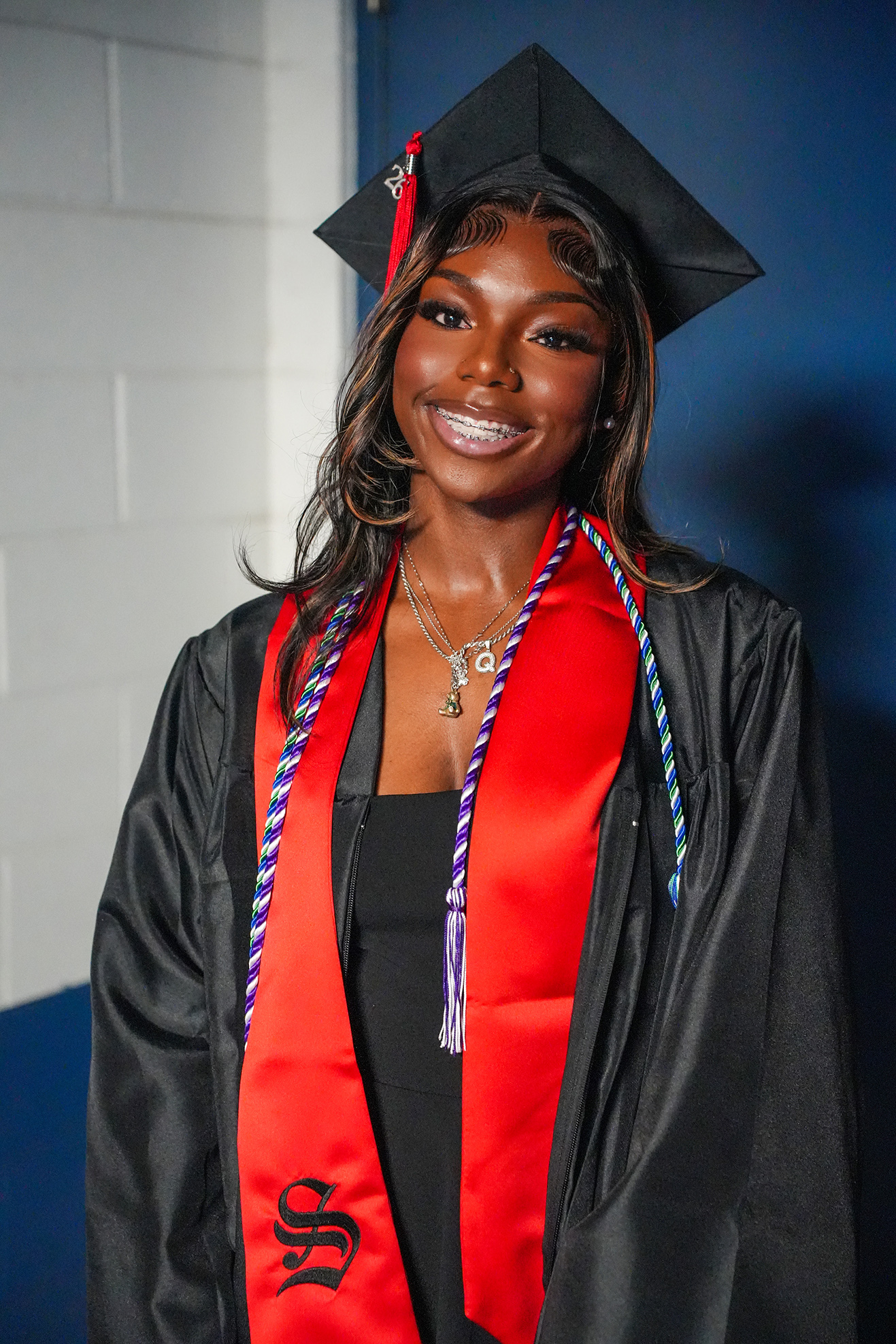A person in a black graduation gown with a red stole and a black graduation cap stands against a white wall.
