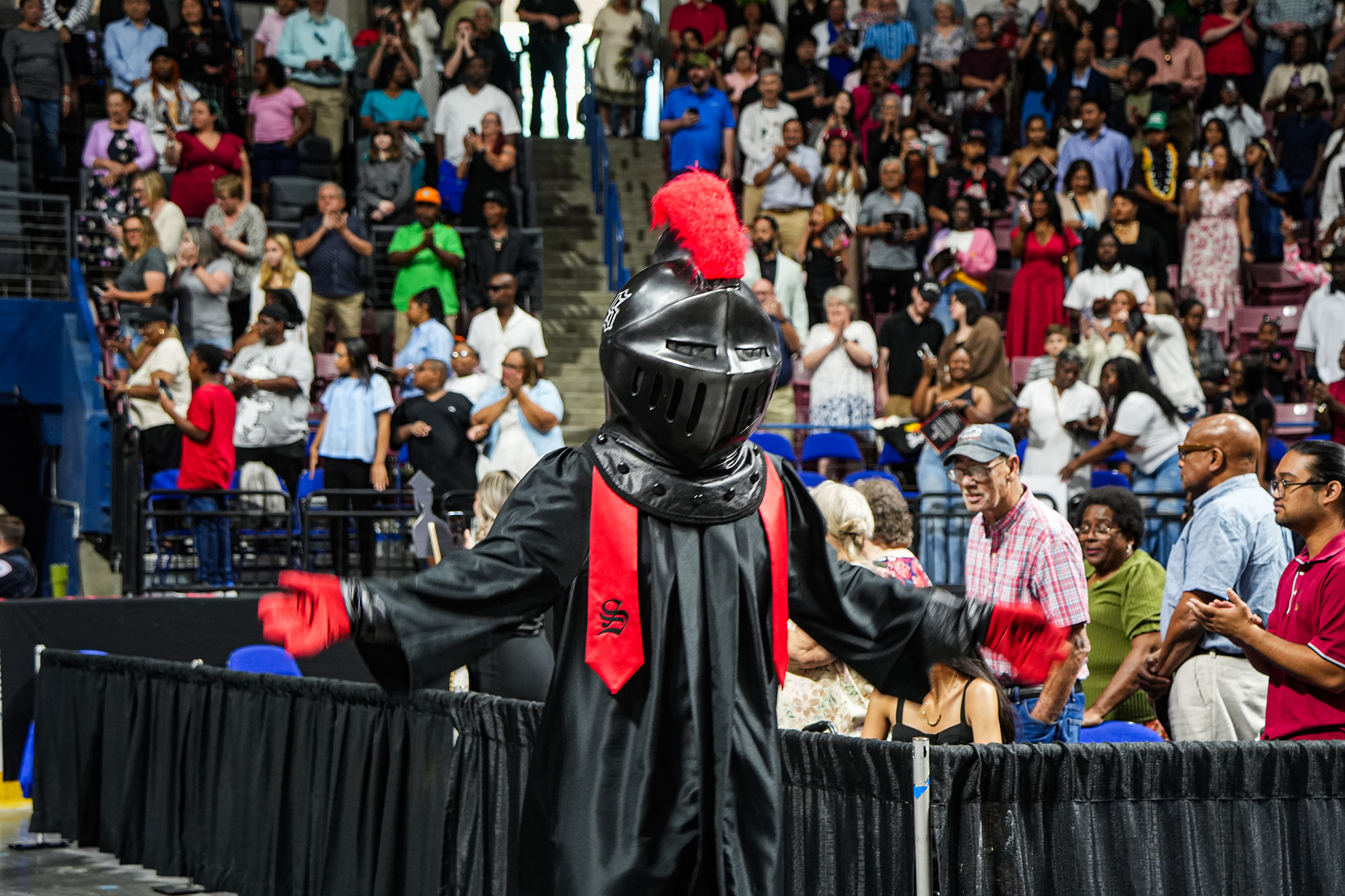 A person in a knight costume stands on stage in front of an audience seated on bleachers.