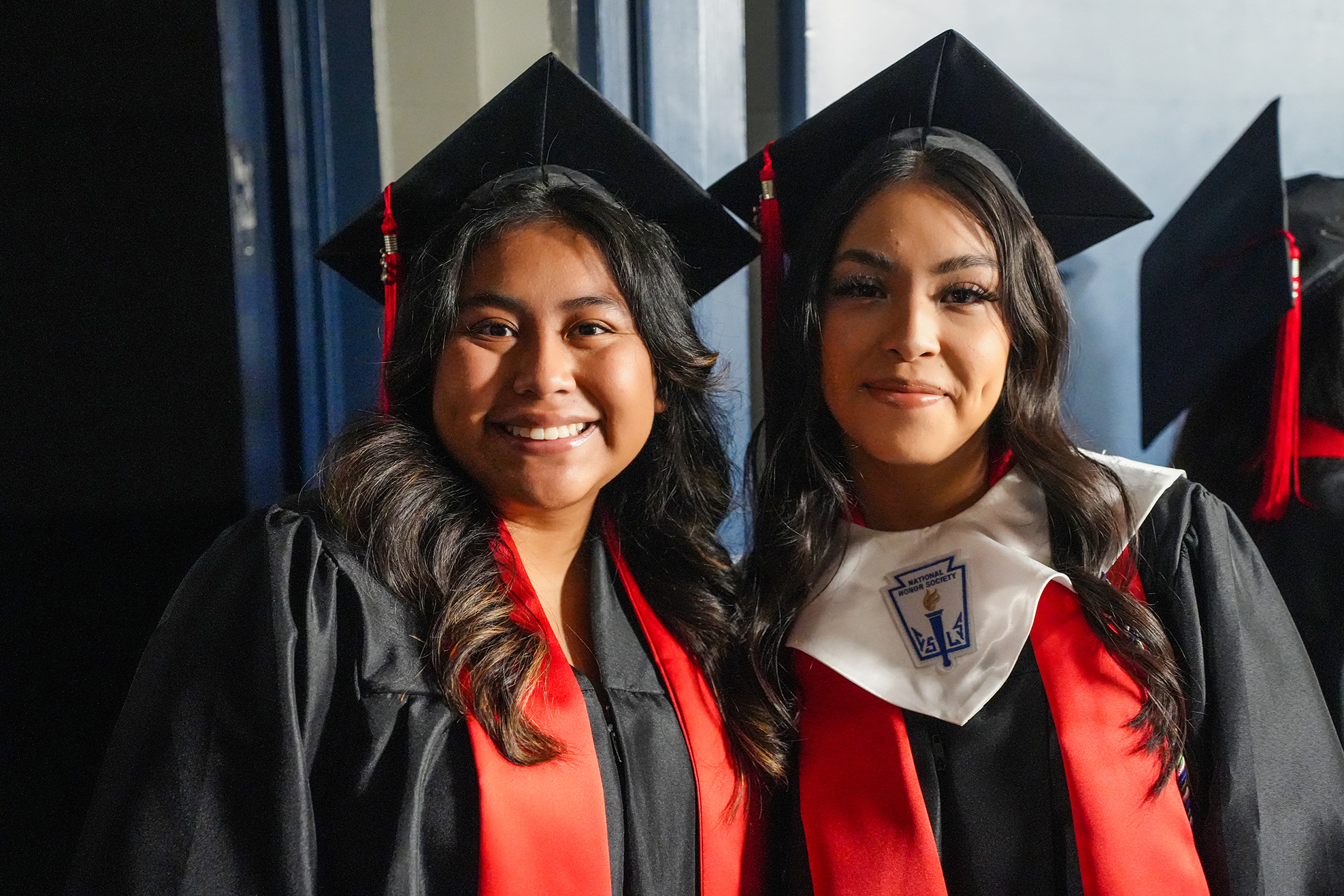 Two women in black graduation gowns with red sashes and caps smile for a photo.