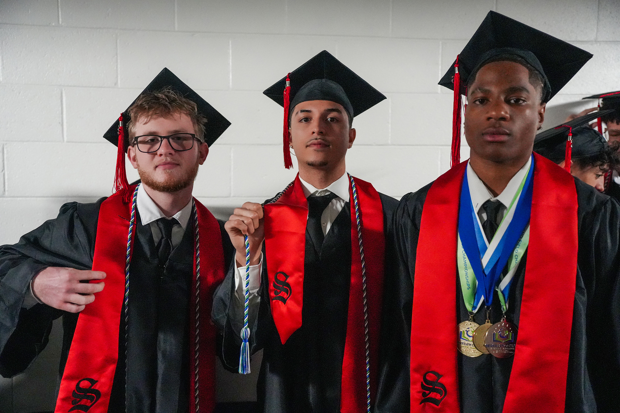 Three men in graduation gowns and caps pose together. They wear red sashes, and one has a medal.